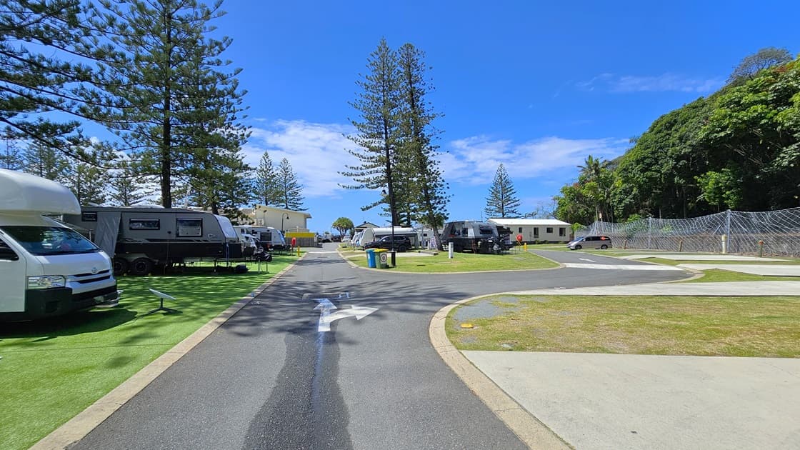 On the left, there are several caravans parked on artificial turfs with vehicles attached to the front of some caravans. Trees line the background,  On the right, there is a paved road with a footpath adjacent to it. More caravans are parked on the grass further along the right side. Tall pine trees stand prominently in the centre of the image separating the left and right sections.  A low-set building is also visible on the far right. The chain fence secures the right.