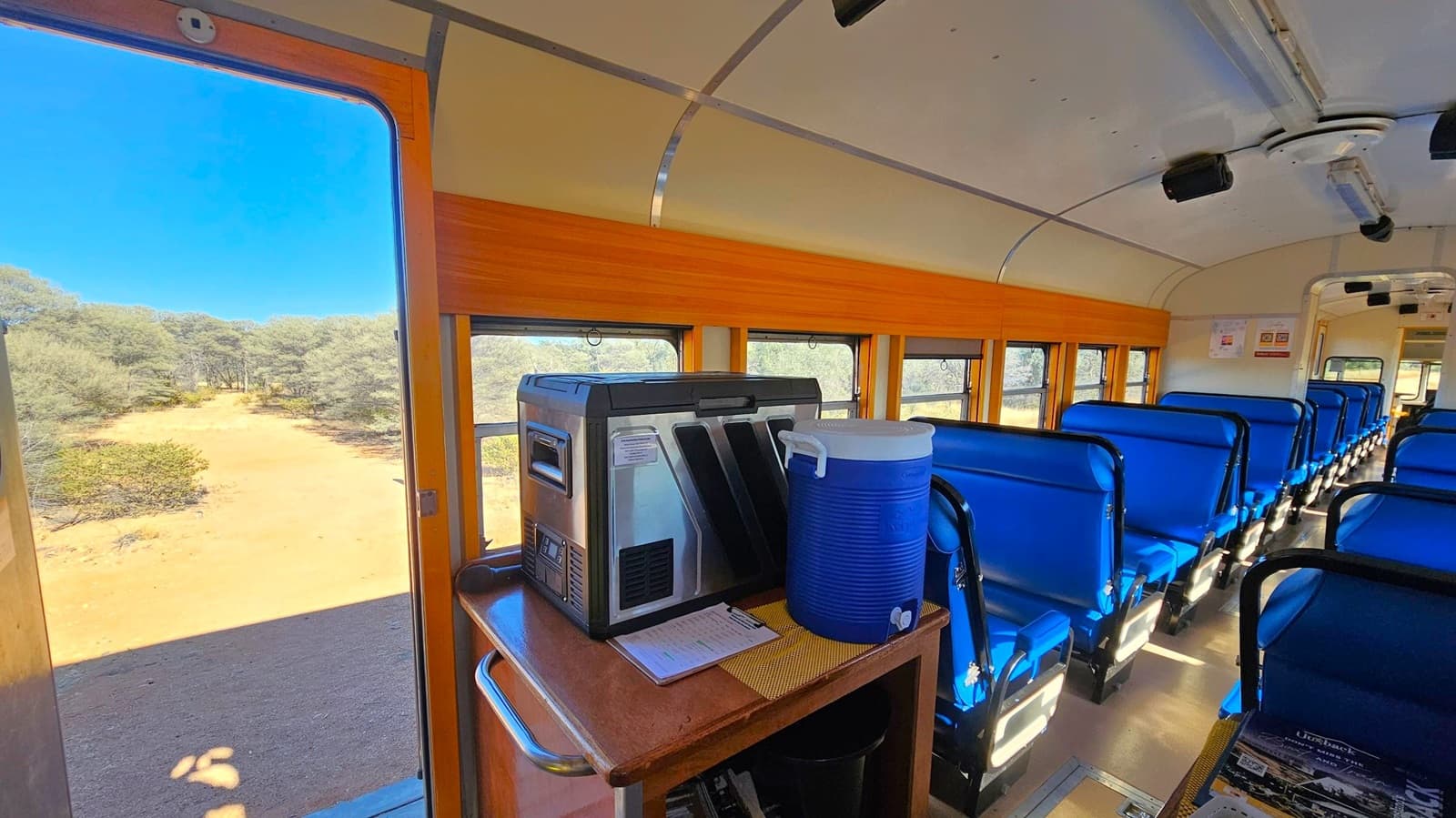 Inside a train carriage with blue seats arranged in rows on the right. A wooden panel runs along the windows. On the left, an open door reveals a view of a sandy path and trees outside. In the foreground, a table holds a large blue cooler and a metallic appliance. The ceiling is curved with lighting fixtures, and the floor is light-coloured. Posters are visible on the far wall.