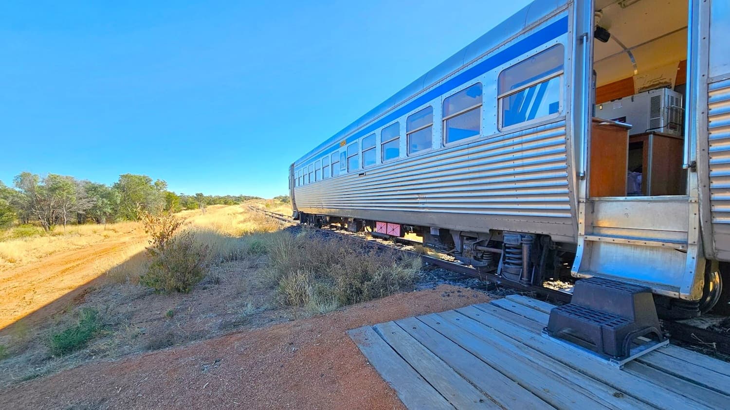 A silver train carriage with blue accents is on the right, with an open door revealing an interior with a desk and air conditioning unit. A small black step is placed on a wooden platform leading to the door. On the left, a dirt path runs alongside the train, bordered by dry grass and scattered bushes. In the background, there are trees and a clear horizon. The ground is a mix of gravel and dirt.