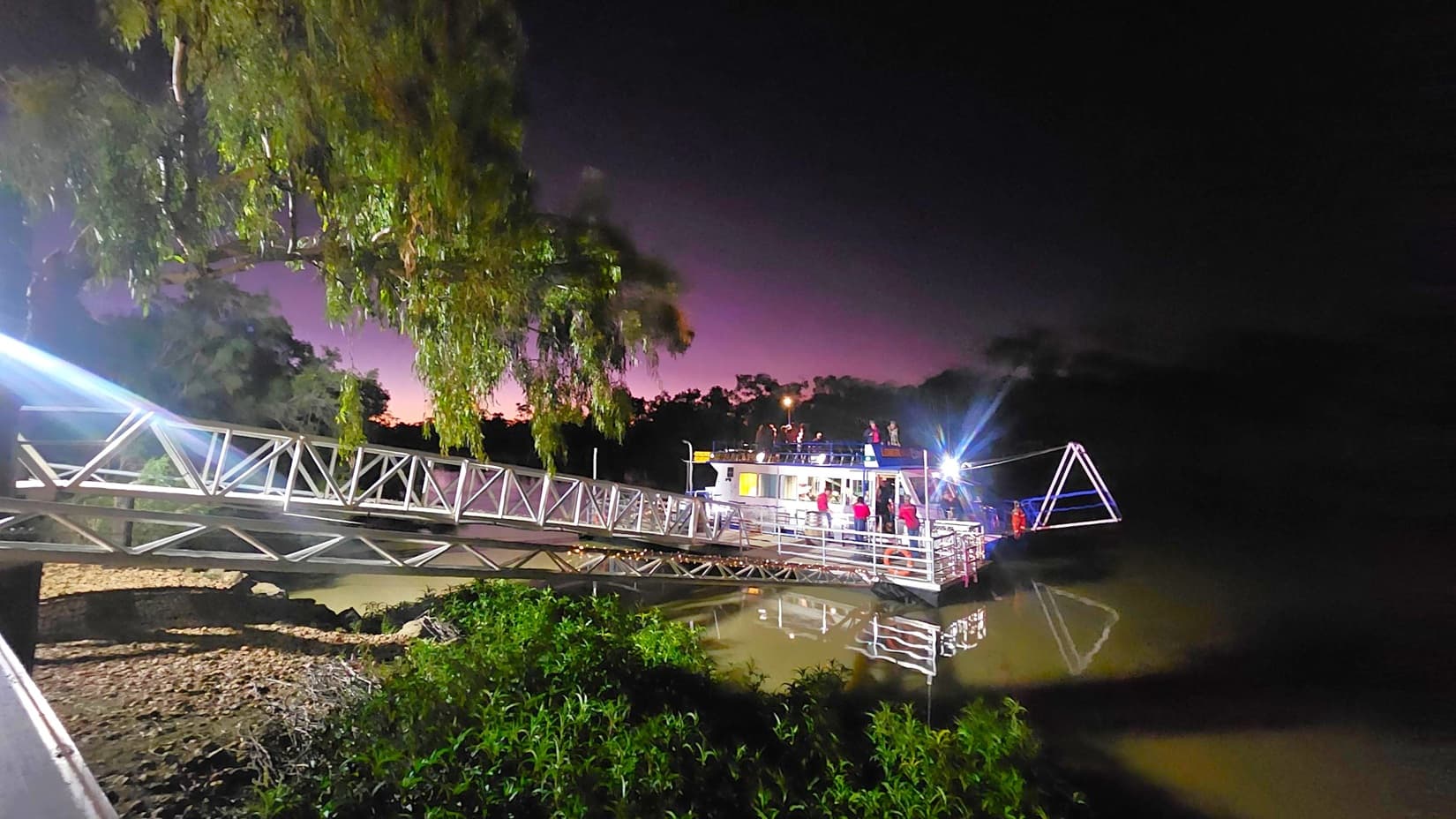 A metal walkway extends from the left, leading to a boat on the right. The boat is illuminated, with people visible on board. Trees with dense foliage are on the left, and the water below reflects the structure and lights. The sky in the background has a gradient from dark to a faint purple hue. Vegetation is visible in the foreground near the water's edge.