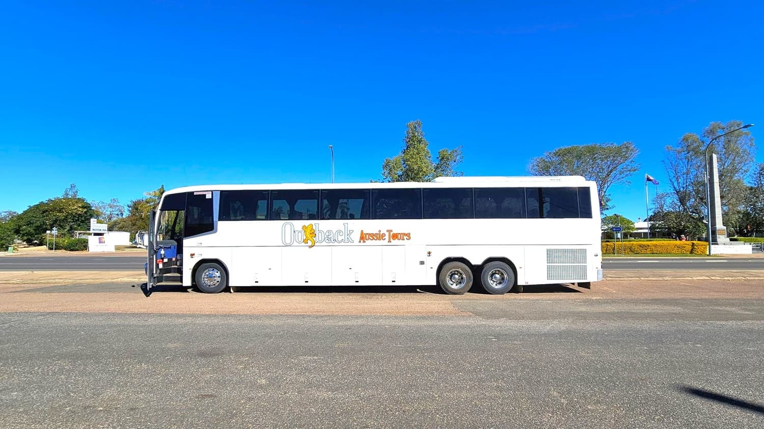 A large white tour bus with "Outback Aussie Tours" written on its side is parked on a paved area. The bus has multiple windows and three axles. In the background, there are trees, a flagpole with a flag, and a tall monument. A sign for a motor inn is visible to the left. The ground is a mix of asphalt and grass, and the sky is clear.