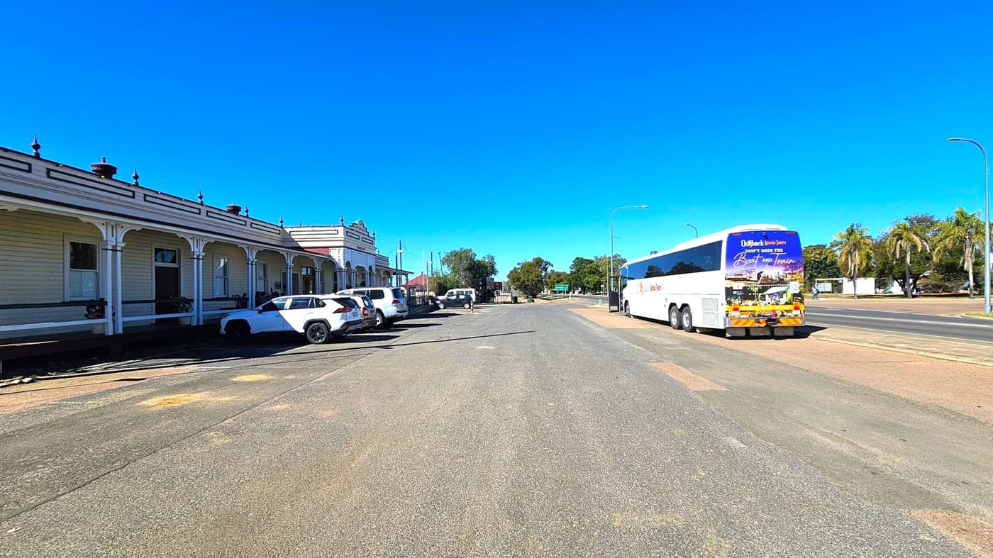 A wide street with a row of parked white cars on the left, adjacent to a long, single-storey building with decorative trim and covered walkways. On the right, a large white coach bus is parked on the road, with palm trees and other greenery in the background. The road surface is a mix of grey and brown patches. The sky is clear and blue.