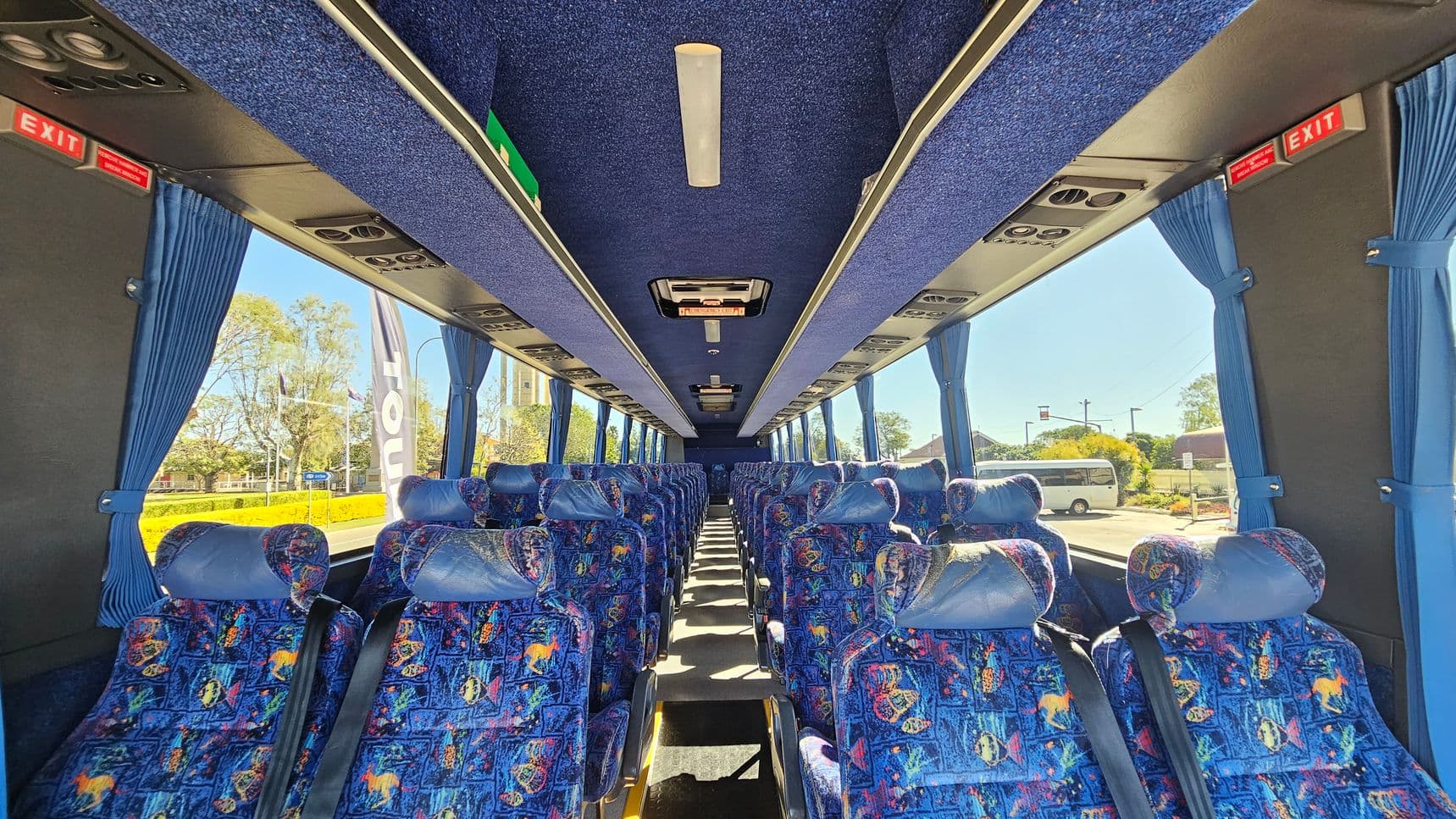 Inside a coach bus, rows of colourful patterned seats with headrests are visible on both sides. Blue curtains hang beside large windows, allowing a view of trees and a parked vehicle outside. The ceiling is lined with blue fabric and features overhead lights and air vents. Red "EXIT" signs are positioned at the front and back. The floor is carpeted in a dark colour, and the aisle runs down the centre.