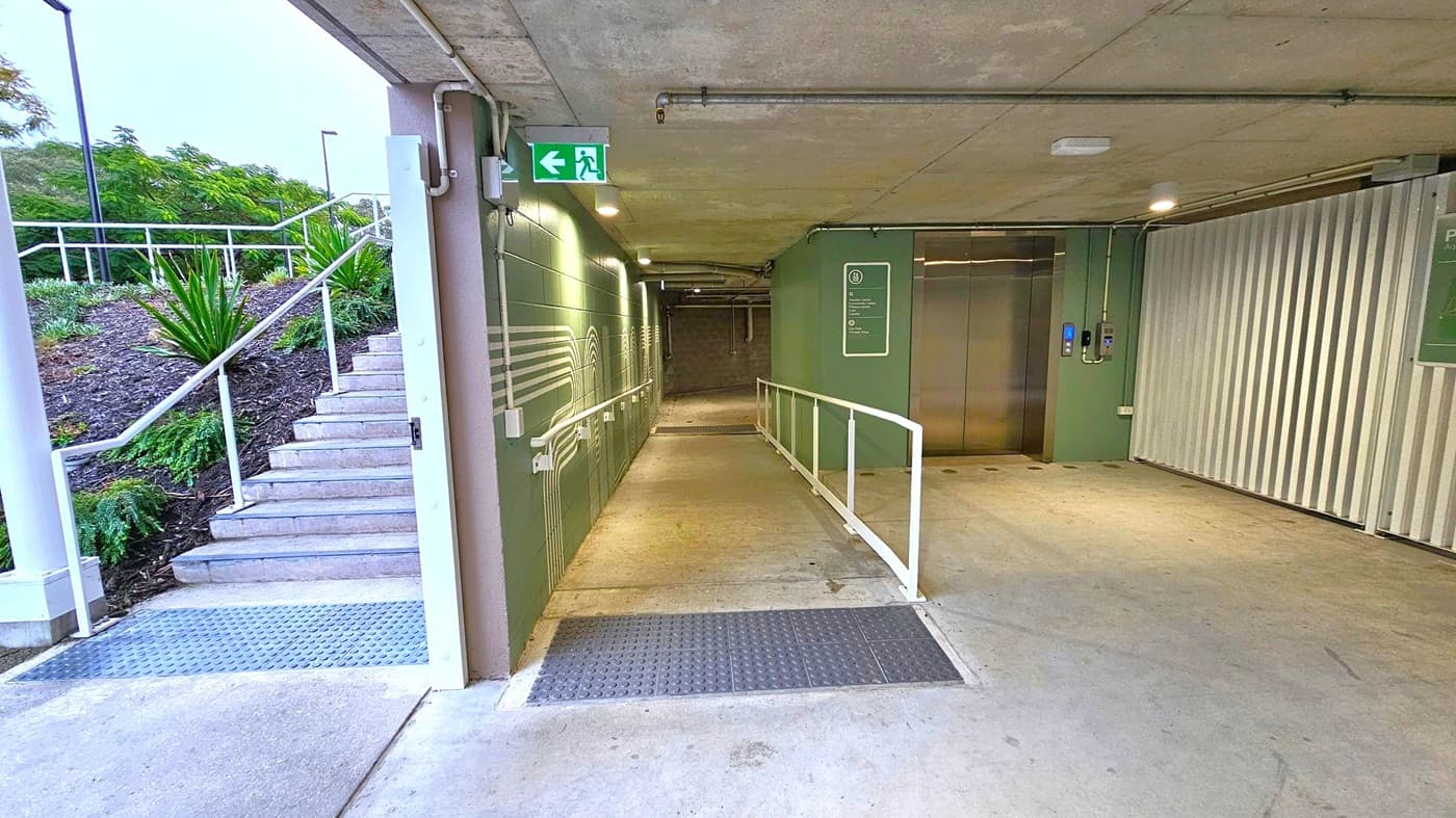 Concrete corridor with a ramp and stairs. On the left, outdoor stairs with white railings lead up, surrounded by greenery. In the middle, a ramp with white railings extends into the corridor, which has green walls with line patterns. An exit sign is above. On the right, a lift with metallic doors is set into the wall, next to a corrugated metal panel. The ceiling is concrete with visible pipes, and the floor is smooth concrete with textured sections.