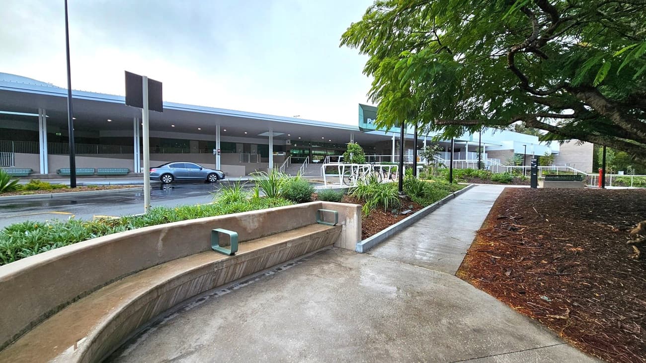 A concrete pathway leads to a building with a covered entrance in the background. On the left, a curved concrete bench is surrounded by greenery. A parked car is visible on the road beside the building. On the right, a large tree with dense foliage provides shade over a mulched area. The building features multiple entrances with steps and railings, and there are several lamp posts along the path. The ground is wet, suggesting recent rain.