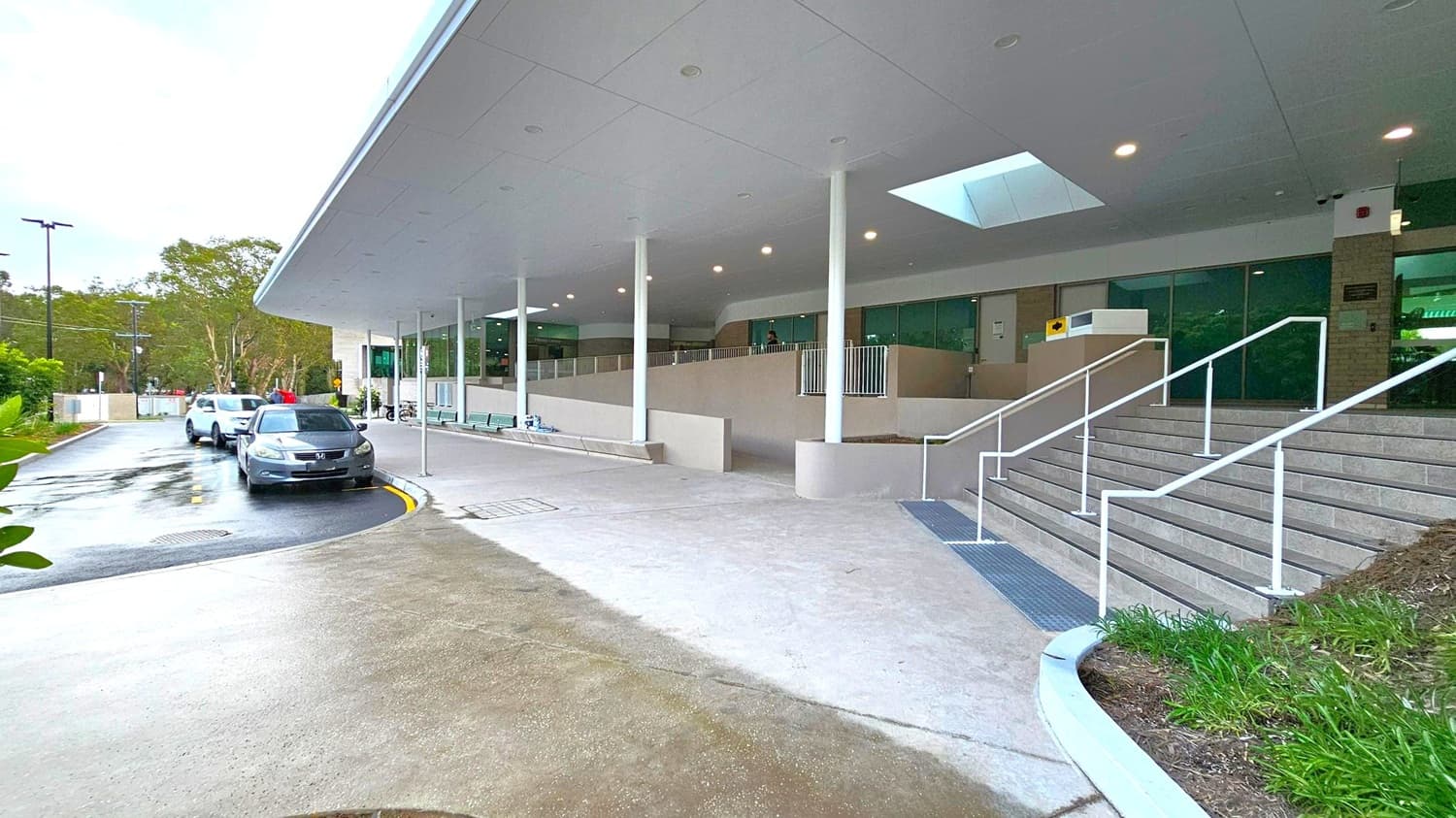 A modern building entrance with a wide, covered walkway supported by white pillars. On the right, a set of stairs with white railings leads up to the entrance. The foreground features a concrete path with some grass on the right. On the left, a driveway curves with parked cars, including a black and a white vehicle. The background shows trees and a cloudy sky. The ceiling is smooth with recessed lighting and a skylight.