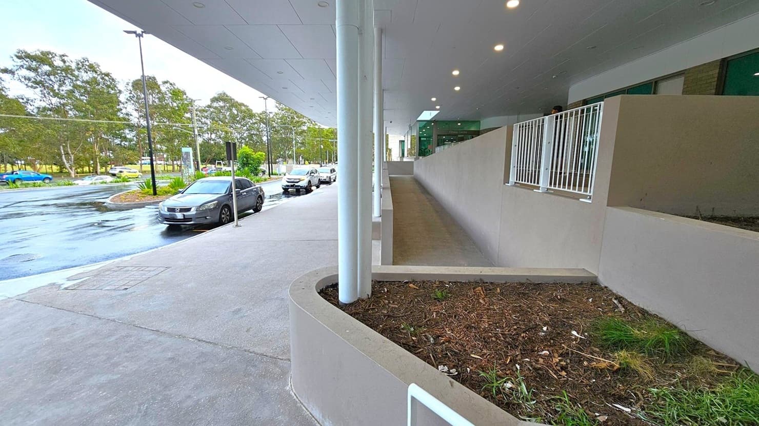 On the left, there is a row of parked cars along a wet road, with trees and a grassy area in the background. On the right, a building with a covered walkway features a sloped ramp and a white railing. The foreground has a small garden bed with mulch and sparse plants. The ceiling above the walkway is lined with recessed lights.