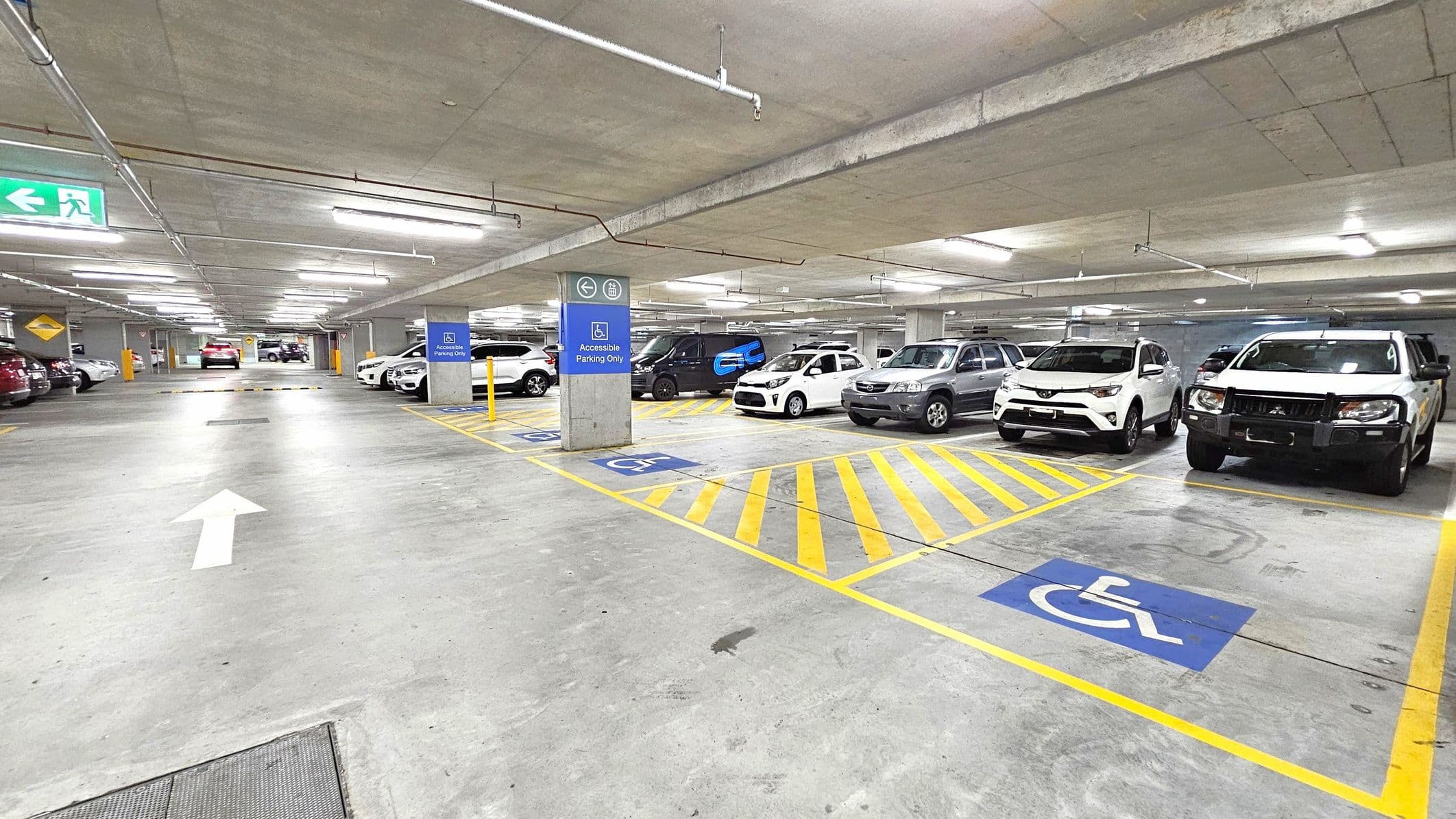 Underground car park with concrete floor and ceiling. On the right, several vehicles are parked in designated accessible parking spaces, marked with blue signs and yellow lines. In the middle, a white arrow is painted on the floor indicating the direction of traffic. On the left, more cars are parked, and a green exit sign is visible. Fluorescent lights are mounted on the ceiling, providing illumination.