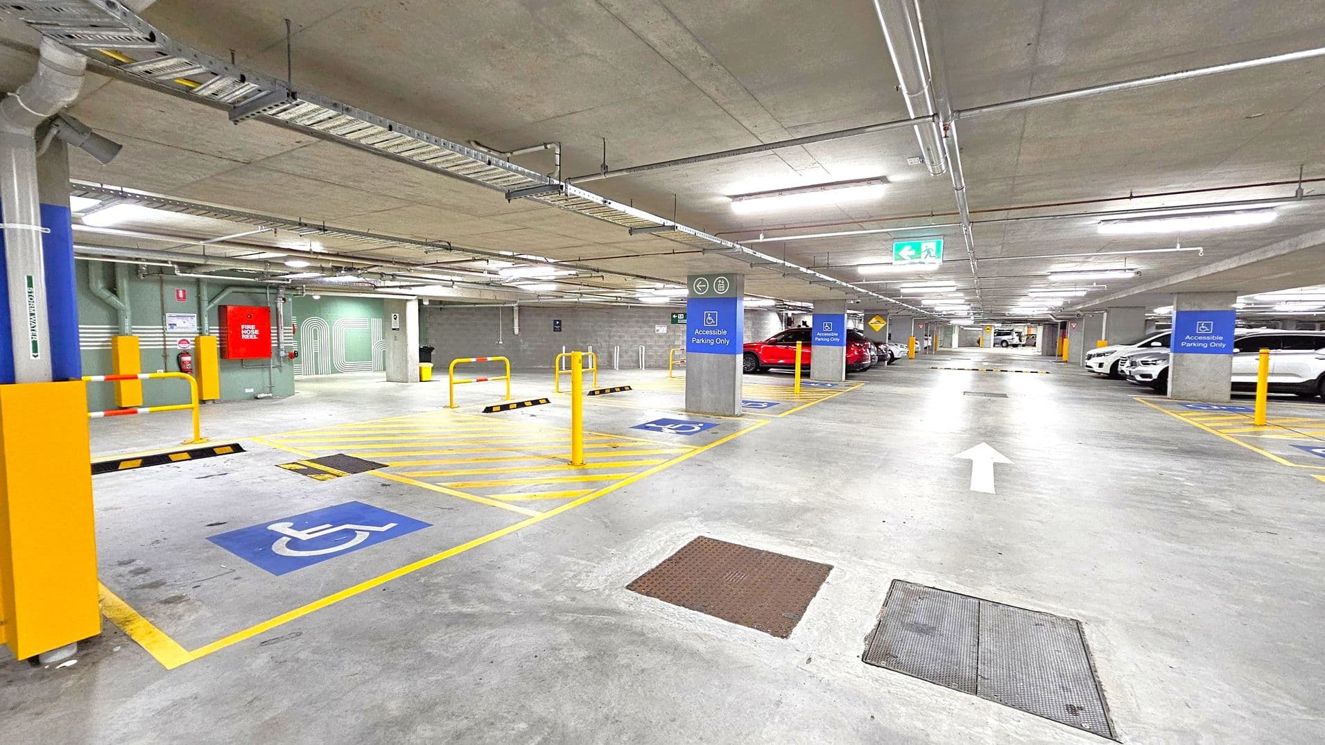 A well-lit underground car park with a concrete floor and ceiling. On the left, there are designated accessible parking spaces marked with blue signs and yellow lines. Yellow bollards and barriers are present near the spaces. On the right, several cars are parked, including a red one. Overhead, there are pipes and fluorescent lights. An arrow on the floor indicates the direction of traffic. In the background, there are more parking spaces and an exit sign.