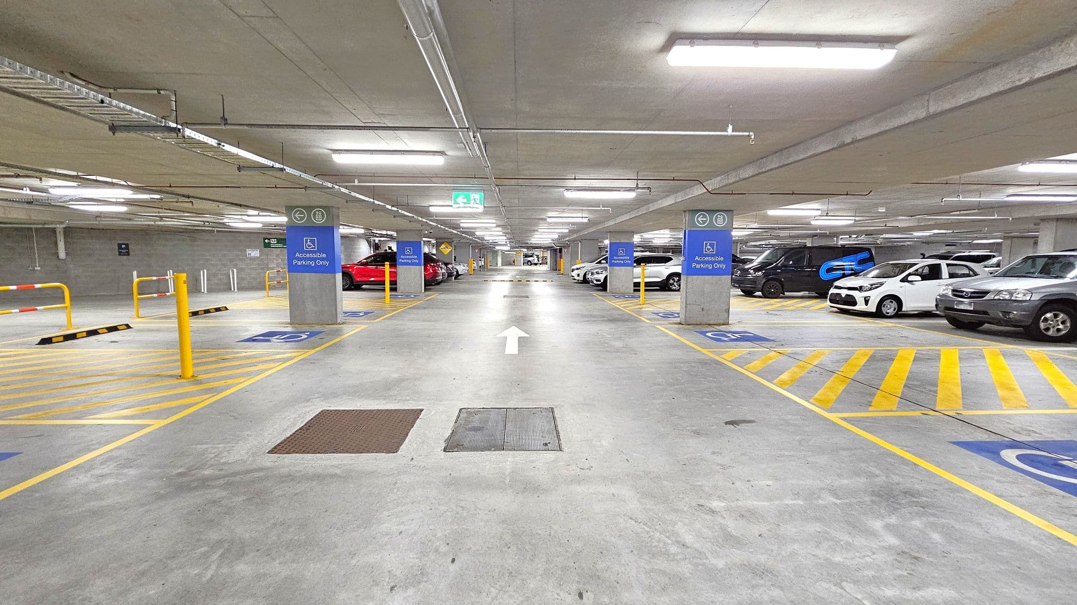 An underground car park with a concrete floor and ceiling. In the foreground, there are accessible parking spaces marked with blue signs and yellow lines. The middle of the image features a wide driving lane with a white arrow indicating direction. On the left and right, various cars are parked, including a red car on the left and a white car on the right. The ceiling has fluorescent lighting and visible pipes. Yellow bollards and barriers are present near the parking spaces.