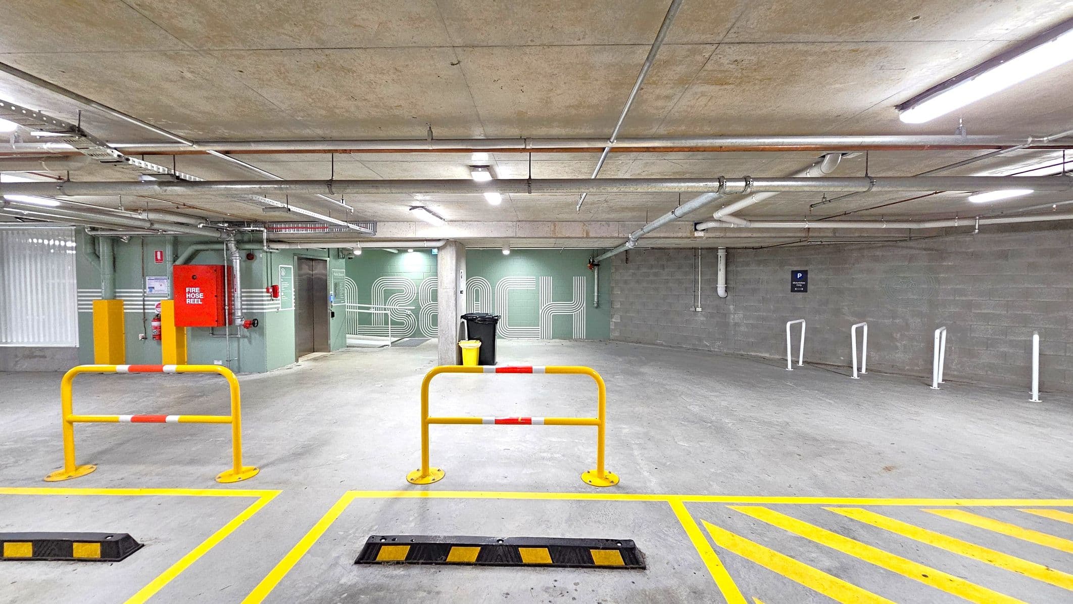 A concrete underground car park with a grey floor and ceiling. In the foreground, yellow and black speed bumps and yellow barriers with red and white stripes are visible. The left side features a red fire hose reel mounted on a green wall, with pipes and a lift nearby. The right side has a grey brick wall with white bike racks and a parking sign. Fluorescent lights and exposed pipes run across the ceiling.