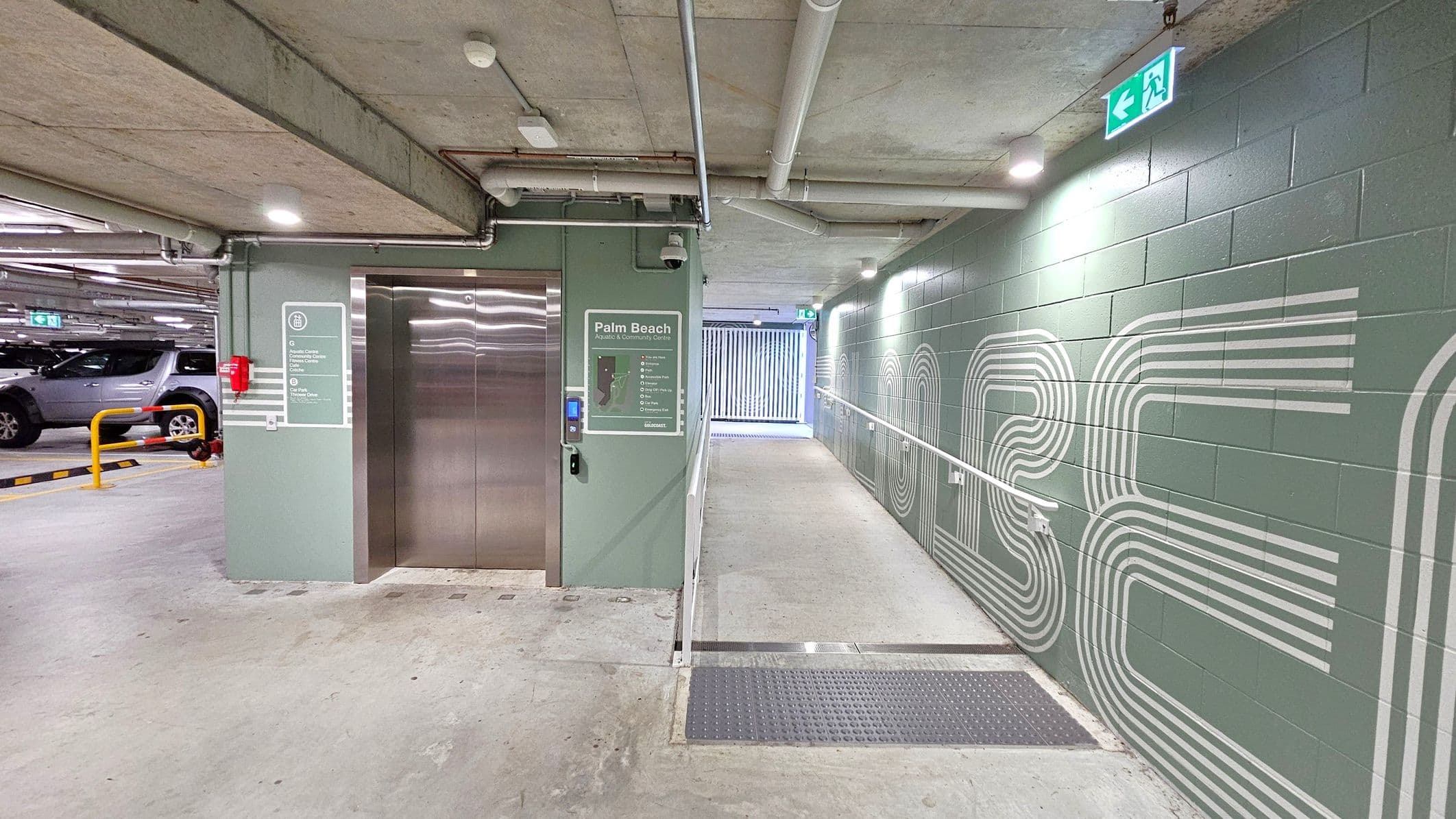 A car park with a concrete floor and ceiling. On the left, there's a lift with a green sign labelled "Palm Beach" and a fire extinguisher nearby. A grey car is parked next to a yellow barrier. On the right, a green wall features white geometric patterns and a lit exit sign. A ramp with a handrail leads to a gated area in the background. Pipes and lights are visible on the ceiling.