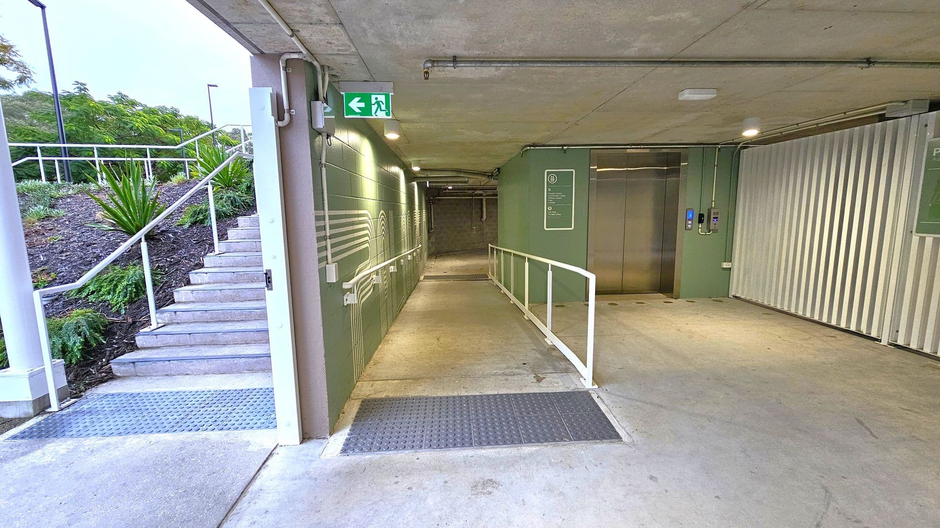A concrete corridor with a ramp and handrails on the right, leading to a lift with metallic doors. On the left, a staircase with white railings ascends to an outdoor area with greenery and small plants. The ceiling is concrete with exposed pipes and lights. An emergency exit sign is visible above the corridor. The floor is smooth concrete with a textured metal section near the entrance.