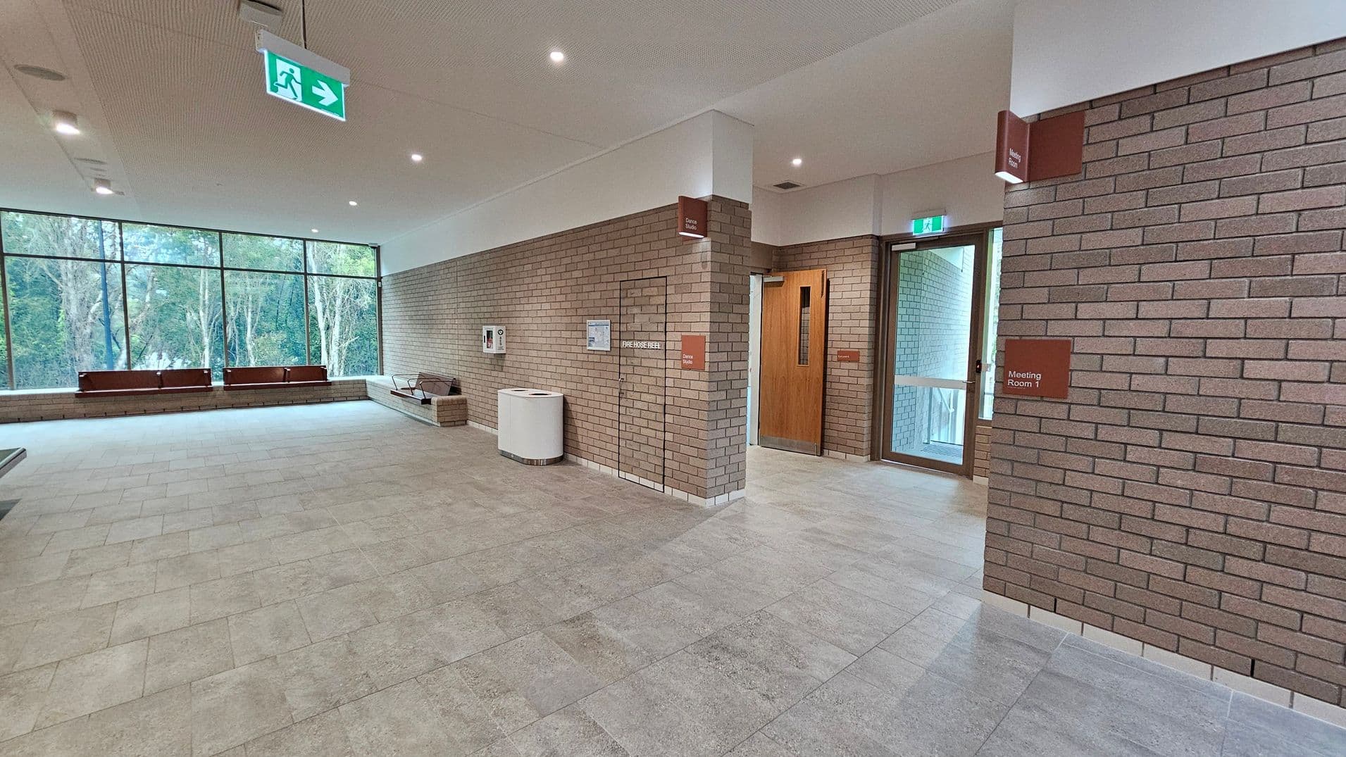 A spacious interior with brick walls and a tiled floor. On the right, there is a wooden door labelled "Meeting Room 1" and a glass door leading outside. In the centre, a fire hose reel is mounted on the wall. On the left, large windows offer a view of trees outside, with benches placed along the window. The ceiling is white with recessed lighting and an illuminated exit sign.