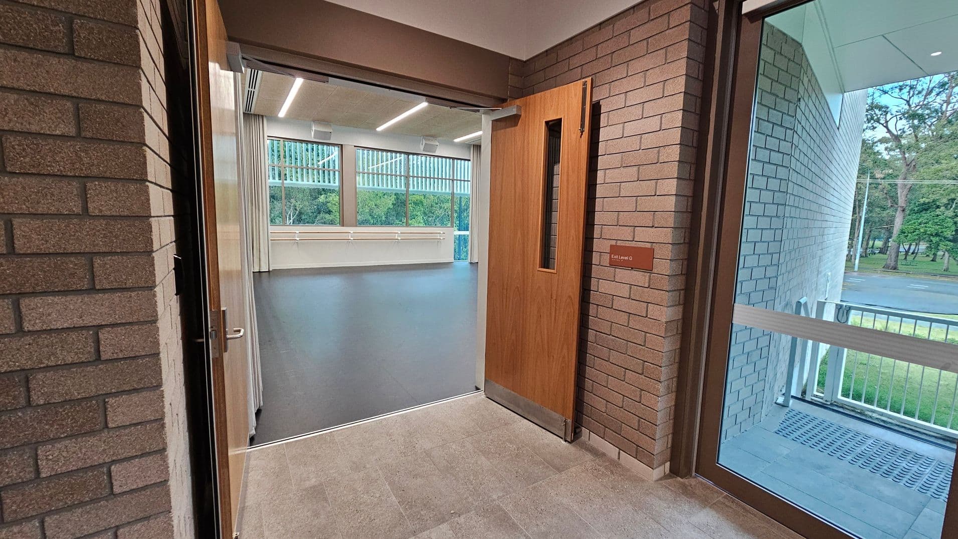 A corridor with brick walls on both sides. On the right, a wooden door with a narrow vertical window is open, leading into a room with a dark floor and large windows showing trees outside. A sign next to the door reads "Exit Level G." The floor in the corridor is tiled, and the ceiling has recessed lighting. On the far right, a glass door opens to an outdoor area with a metal grate and more trees visible.