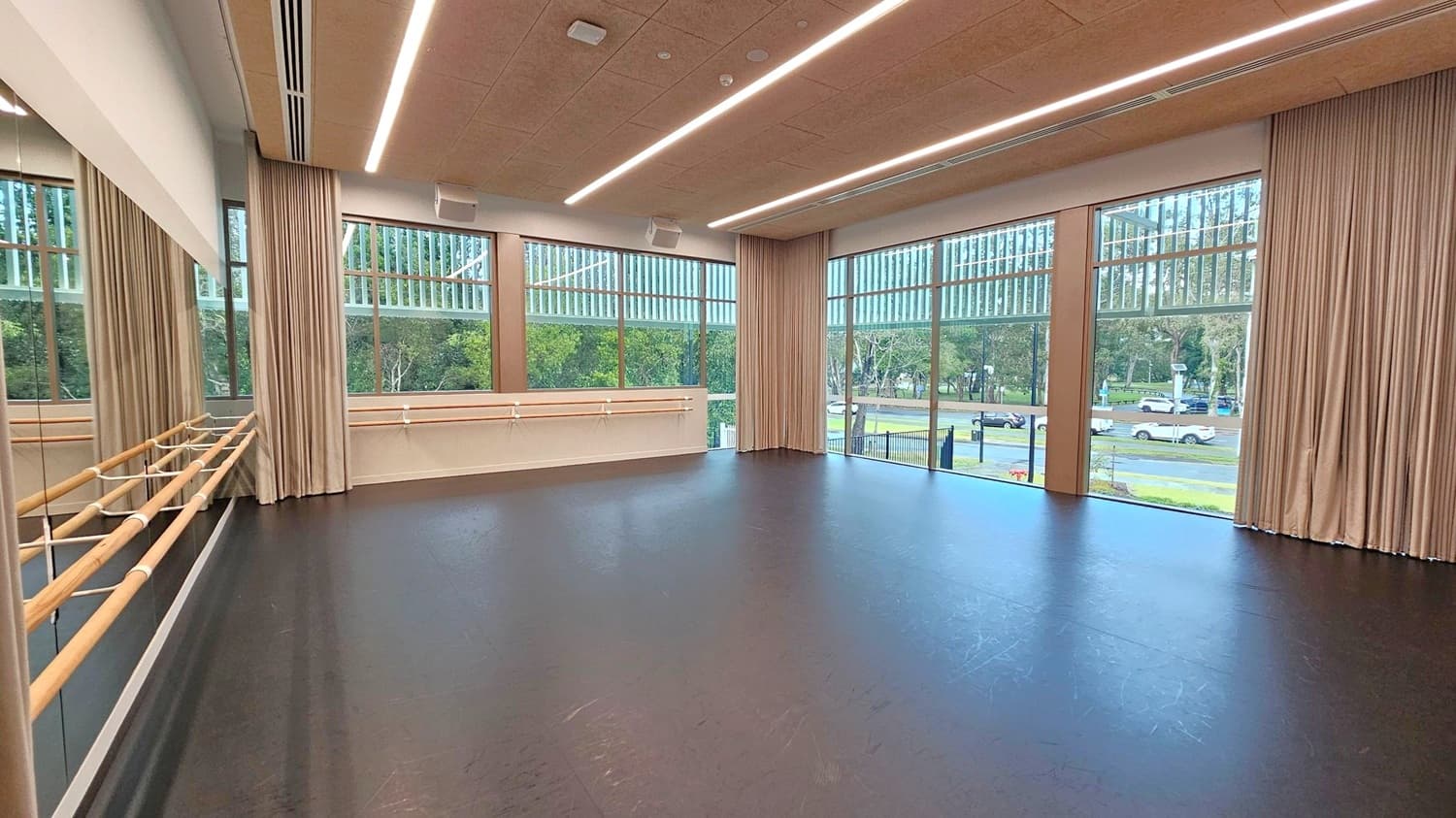 A spacious dance studio with a dark, polished floor and a high ceiling featuring linear lights. On the left, there is a wall with large mirrors and wooden ballet barres. The right side has large windows with vertical blinds, offering a view of trees and parked cars outside. Beige curtains frame the windows. The room is well-lit and has a modern, clean design.