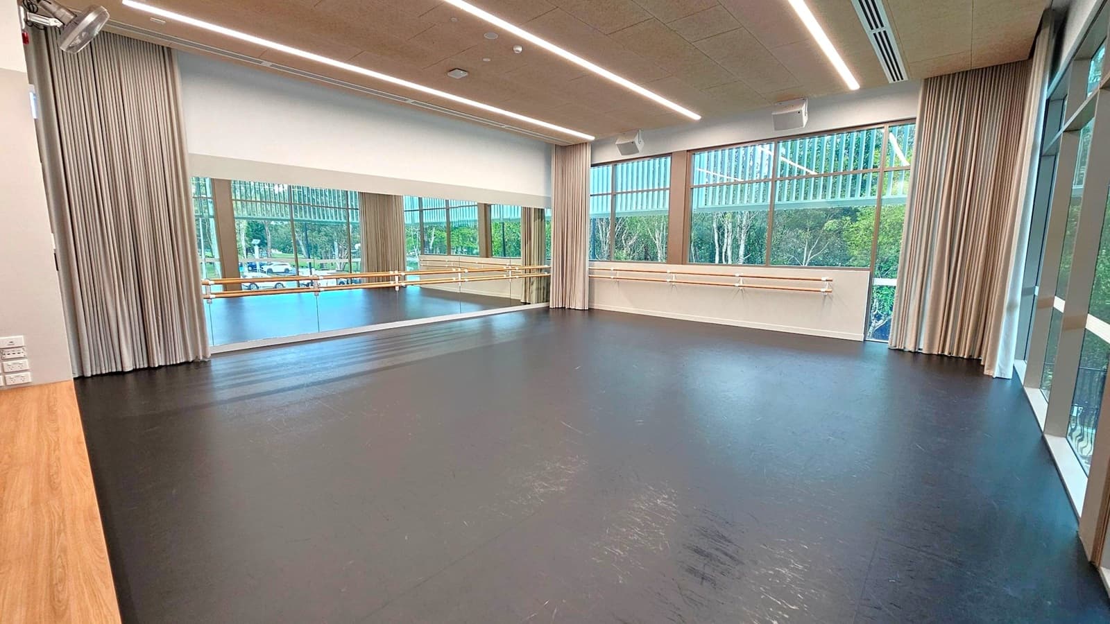 A spacious dance studio with a black floor and a wooden section on the left. The back wall features large mirrors and ballet barres. Tall windows on the right and back walls allow natural light, with views of trees outside. Beige curtains frame the windows. The ceiling is fitted with linear lights and ventilation ducts.