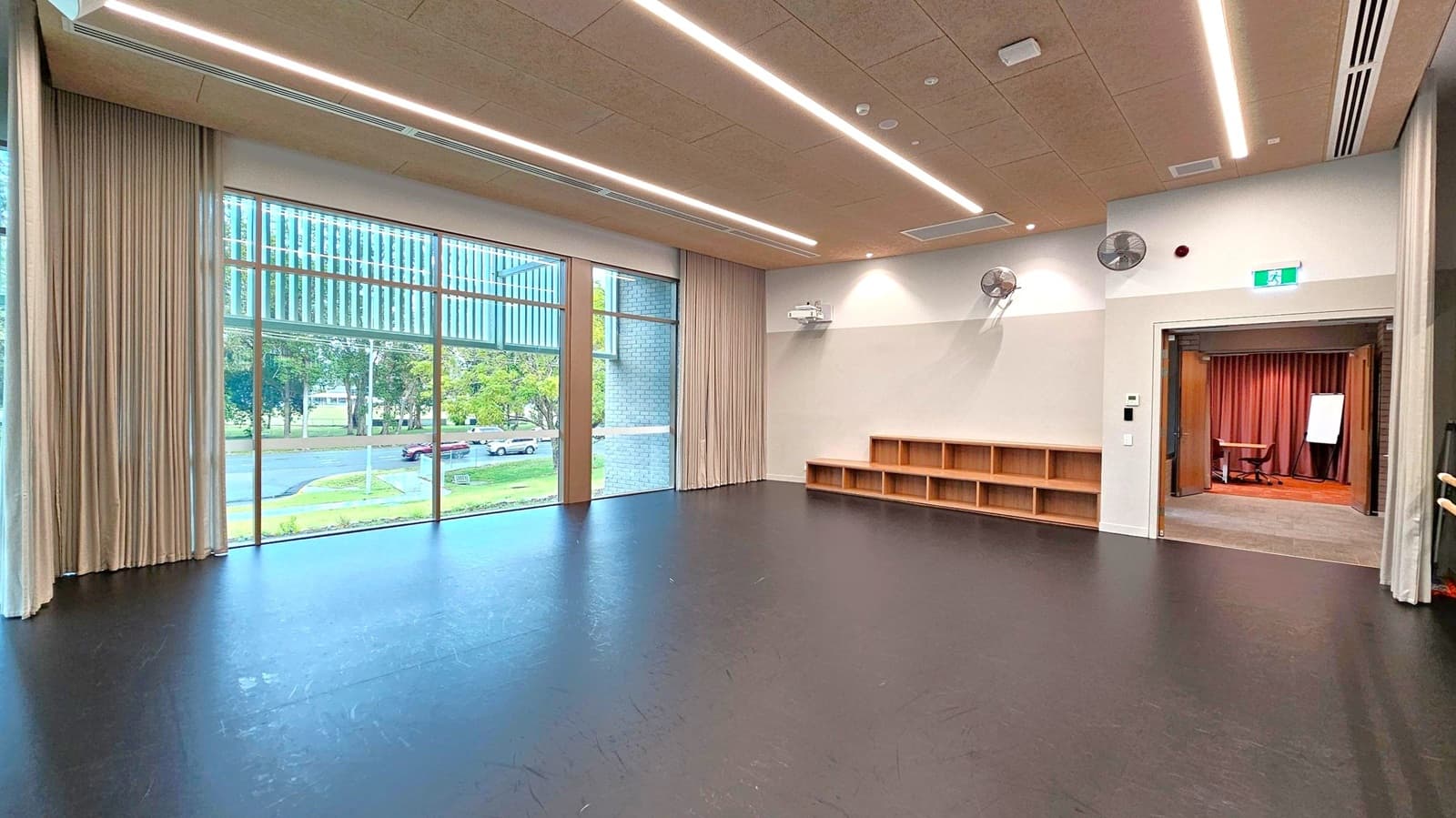 A spacious room with a dark floor and a ceiling featuring linear lights. On the left, large windows with beige curtains offer a view of trees and parked cars outside. The right side has a wall with wooden shelves and two wall-mounted fans. An open doorway leads to another room with a carpeted floor, a table, and a flip chart. An exit sign is above the doorway.