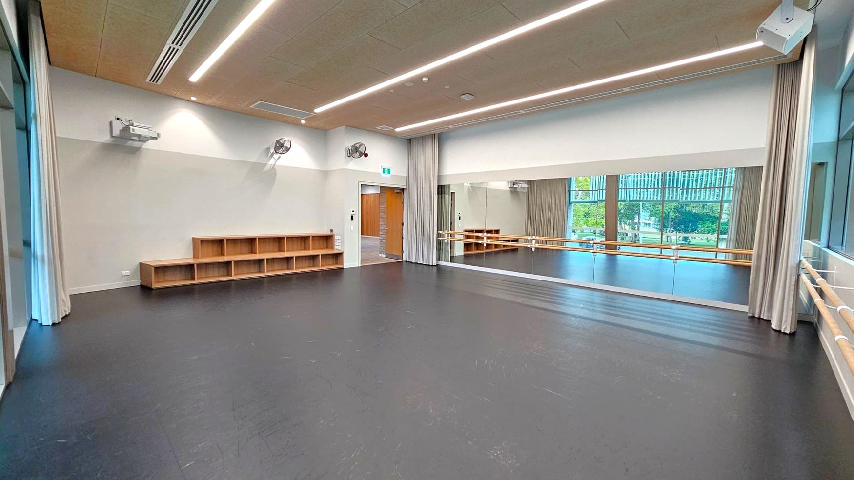 A spacious dance studio with a black floor and a light-coloured ceiling featuring recessed lighting. On the right, there is a large mirror with wooden ballet barres in front. The left side has a wall with wooden cubby shelves and a mounted projector. A door with a curtain is visible near the centre. Large windows in the background provide a view of greenery outside.