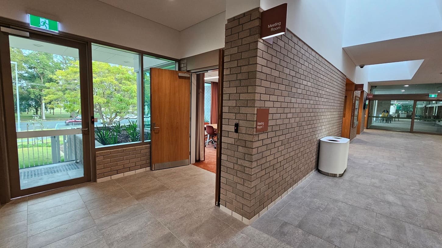 Hallway with grey tiled floor and brick walls. On the left, a glass door with an exit sign above leads outside, showing trees and parked cars. A wooden door is slightly open, revealing a meeting room with chairs. On the right, a sign reads "Meeting Room 1" above a white cylindrical bin. Further down, another glass door is visible, with a glimpse of chairs and tables inside. The ceiling is white with recessed lighting.