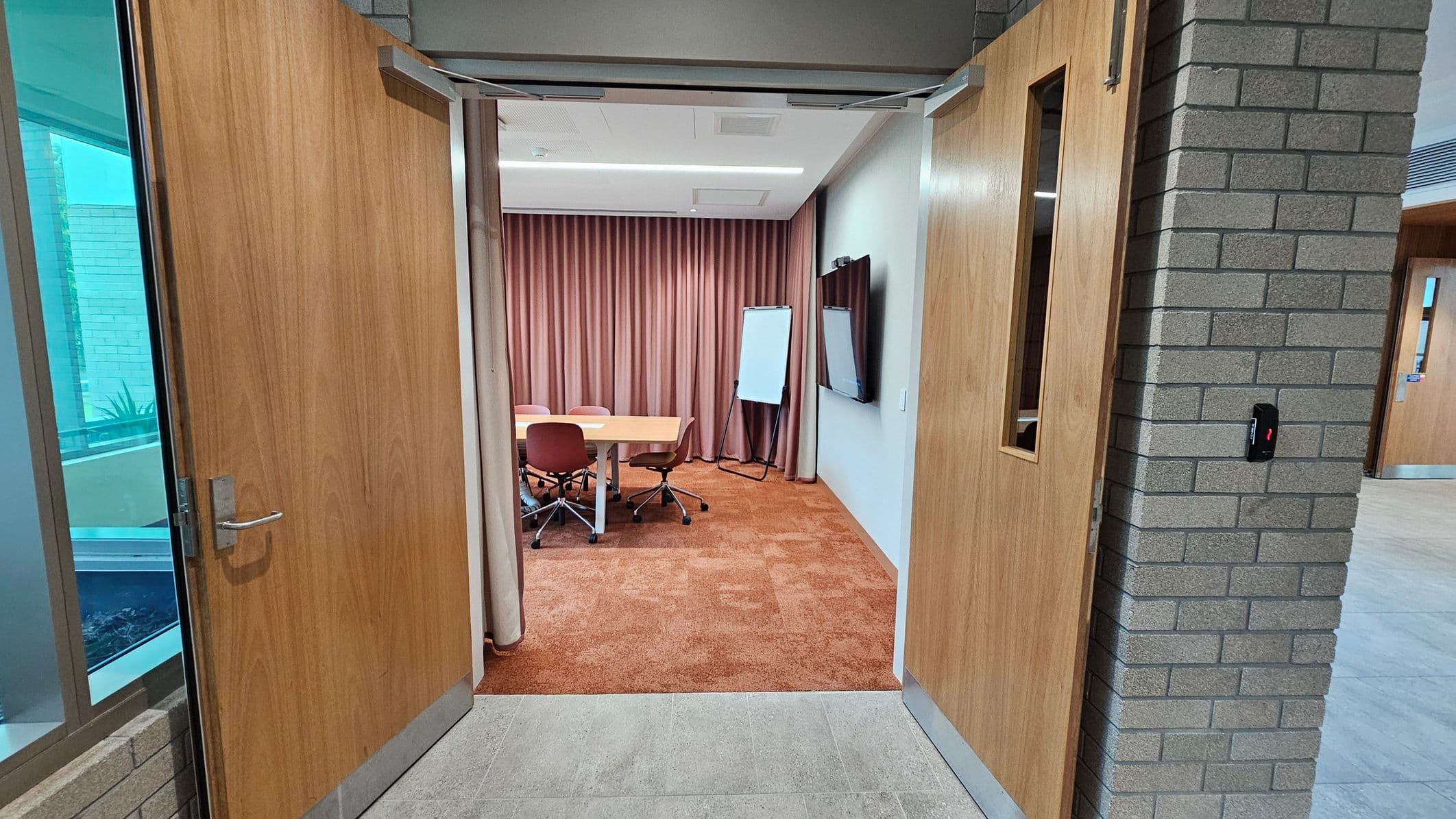 A meeting room with open wooden double doors. Inside, a rectangular table with four brown chairs on wheels is visible. A whiteboard stands on an easel to the right, next to a large wall-mounted screen. The room has brown carpet and is enclosed by floor-to-ceiling curtains. The exterior features a grey brick wall and a window with a view of an outdoor area. The floor outside the room is tiled.