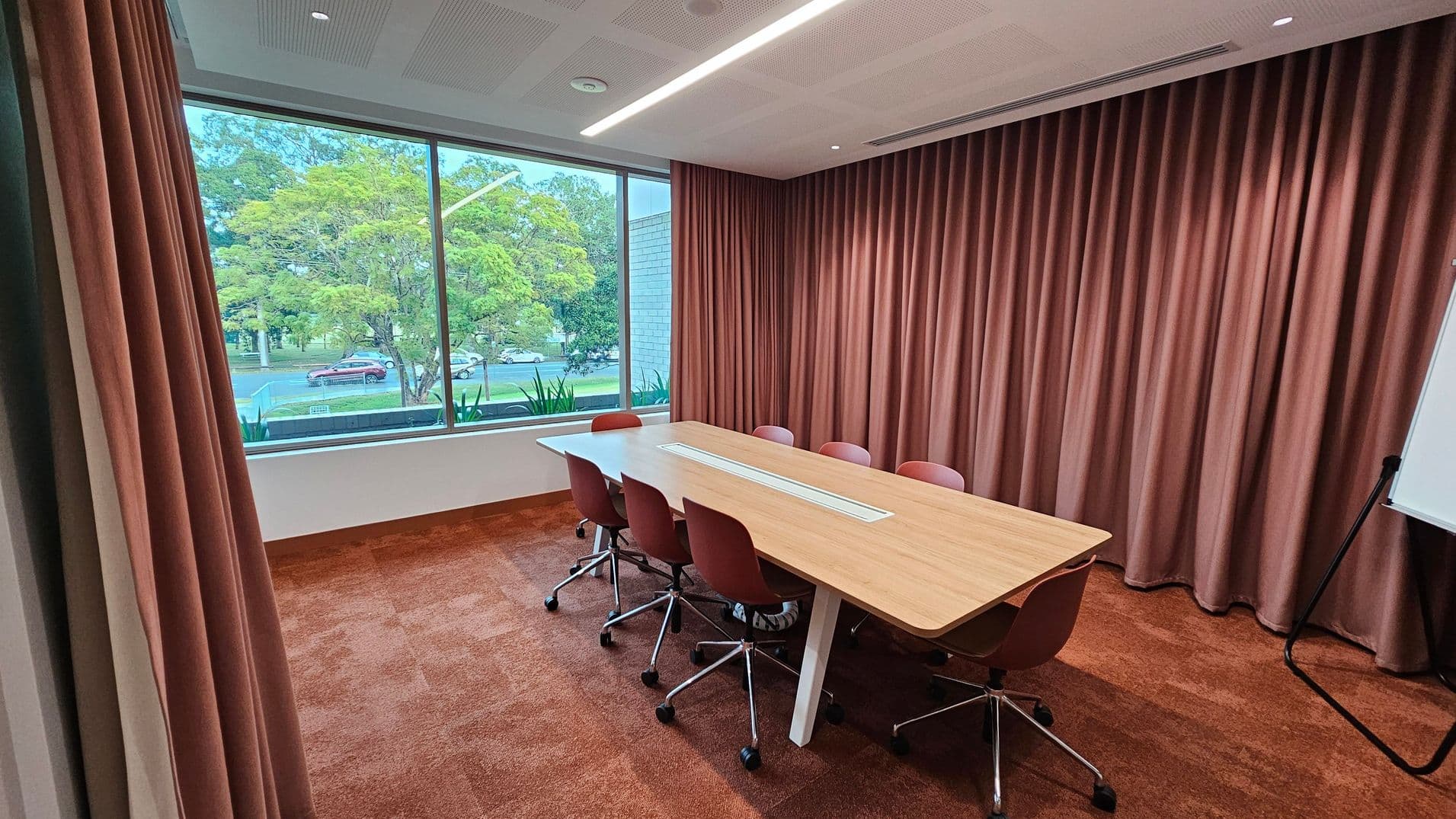 A meeting room with a large window on the left, showing trees and a road outside. The room has a long wooden table with six red chairs on wheels. The right side features floor-to-ceiling red curtains. A whiteboard on a stand is partially visible on the far right. The floor is covered with a red carpet, and the ceiling has recessed lighting and a linear light fixture.
