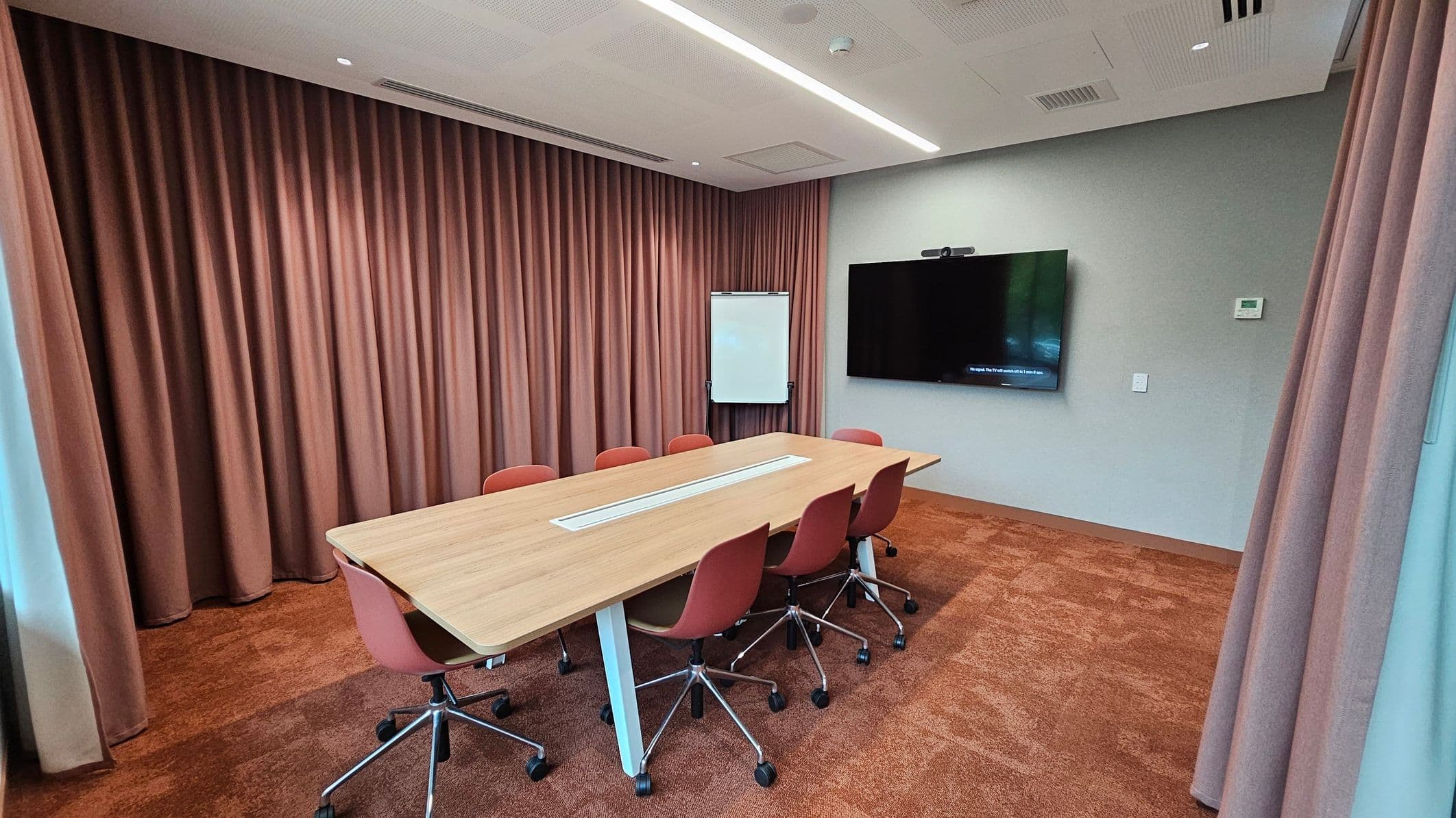 A conference room with a long wooden table surrounded by six red chairs on wheels. The left side features floor-to-ceiling brown curtains. In the centre, a whiteboard stands against the curtain. On the right wall, a large flat-screen TV is mounted, with a thermostat and light switch nearby. The floor is covered in brown carpet, and the ceiling has recessed lighting and ventilation panels.