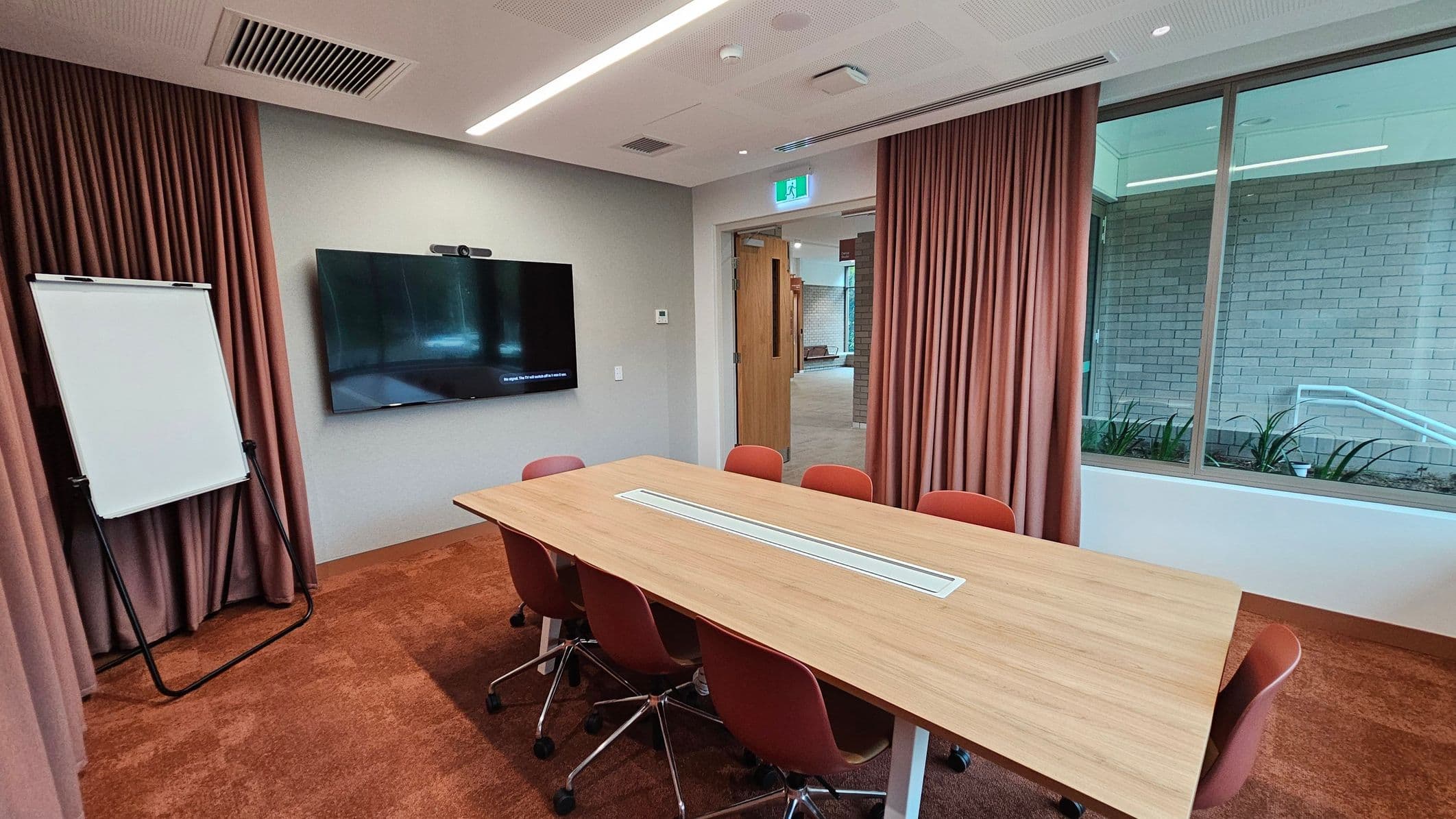 A conference room with a rectangular wooden table surrounded by red chairs on a red carpeted floor. On the left, there's a flip chart and a large wall-mounted screen. The right side features a large window with a view of a brick wall and plants outside. The ceiling has recessed lighting and air vents. A doorway with an open wooden door leads to another area. Brown curtains are drawn along the walls.