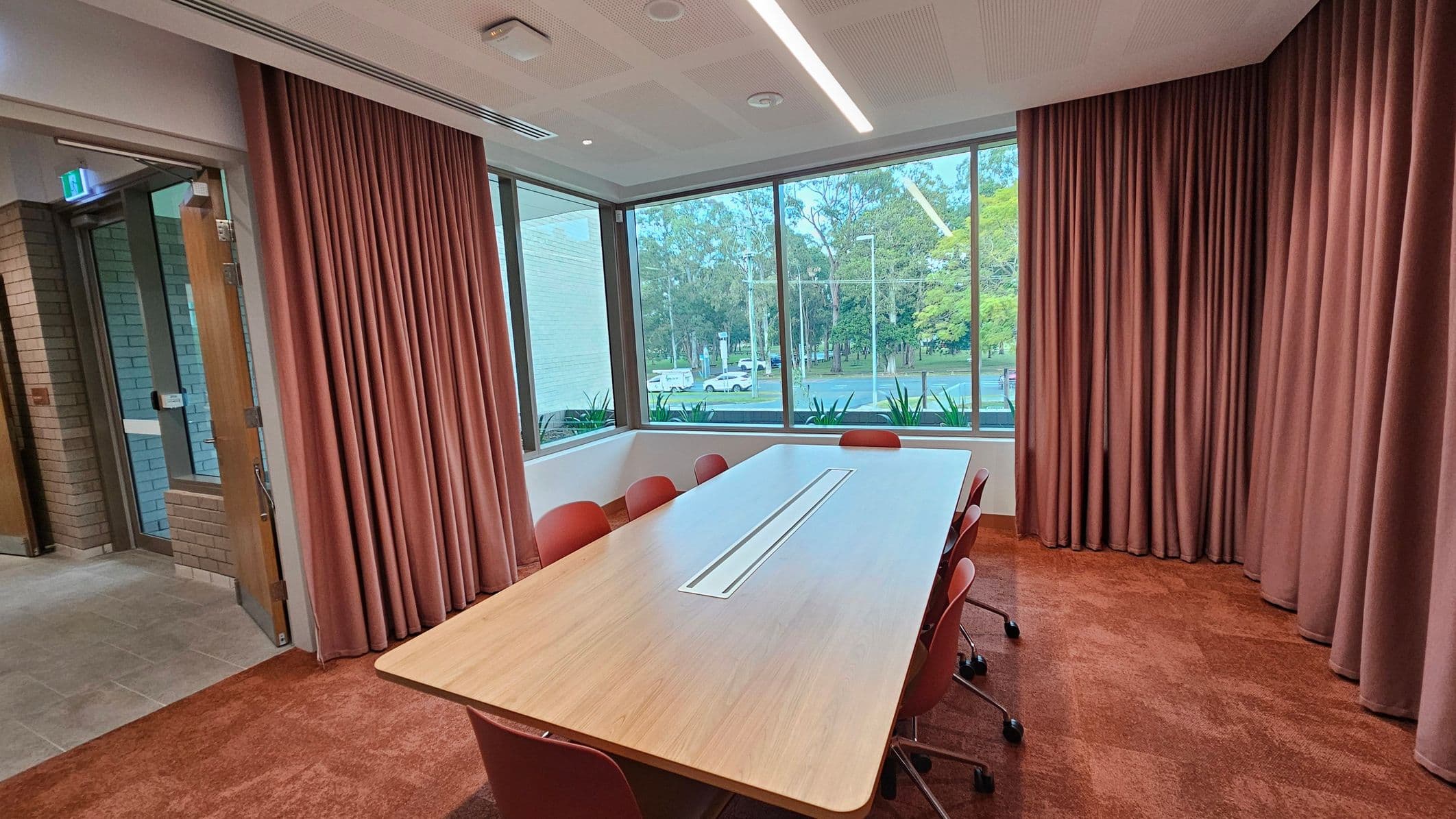 A conference room with a long rectangular table in the centre, surrounded by red chairs on a red carpeted floor. Large windows on the right provide a view of trees and a road outside. The room is enclosed by pinkish curtains. On the left, there's a glass door leading to a hallway with tiled flooring. The ceiling features recessed lighting and ventilation panels.