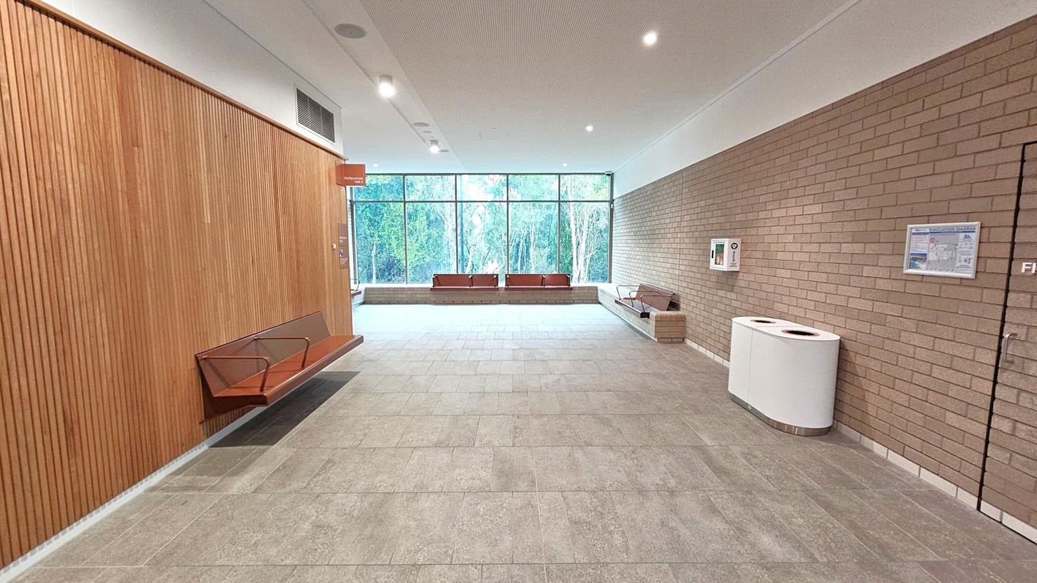 A spacious interior with a tiled floor and a ceiling with recessed lighting. On the left, a wooden panelled wall features a bench with metal armrests. The right wall is made of light brown bricks, with a white bin and some notices. In the centre, large windows offer a view of trees outside. At the far end, more benches are positioned against the window, and a small sign hangs from the ceiling near the wooden wall.