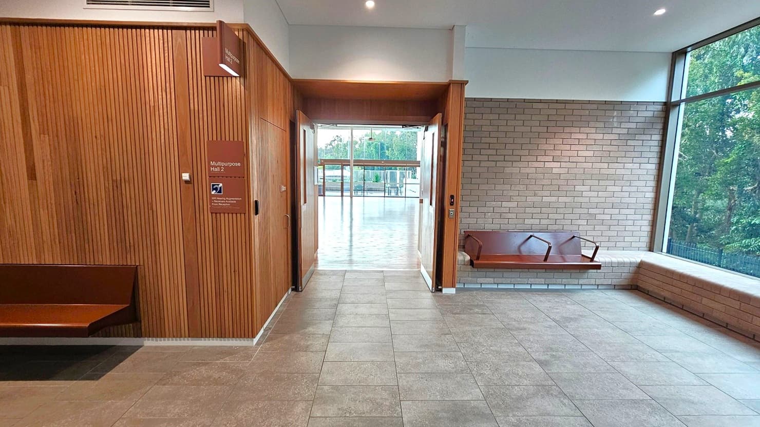 A hallway with a tiled floor and a wooden panelled wall on the left, featuring a sign for "Multipurpose Hall 2". On the right, a brick wall with a built-in bench and large windows showing trees outside. In the centre, a doorway leads to a room with a wooden floor and more windows, allowing natural light to enter. The ceiling is white with recessed lighting.