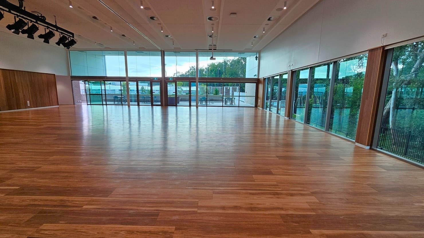 A spacious room with a polished wooden floor and a high ceiling. The left and right walls are lined with large glass windows, allowing natural light to flood in and providing views of trees and outdoor seating. The ceiling features recessed lighting and a row of stage lights on the left. The back wall is mostly glass with doors leading outside, and there are wooden panels on the walls.