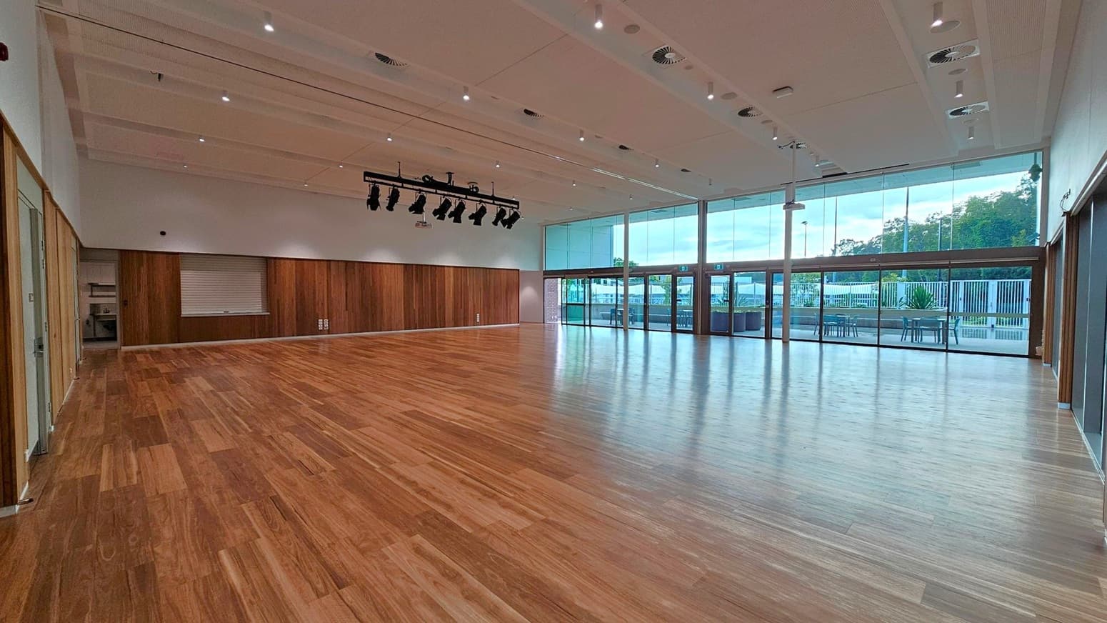 A spacious room with a polished wooden floor and a high ceiling featuring recessed lighting and air vents. On the left, there are wooden panelled walls with a small window and a door. In the centre, a set of stage lights is suspended from the ceiling. The right side has large glass windows and doors leading to an outdoor area with tables and chairs. The background shows greenery and a fence outside.
