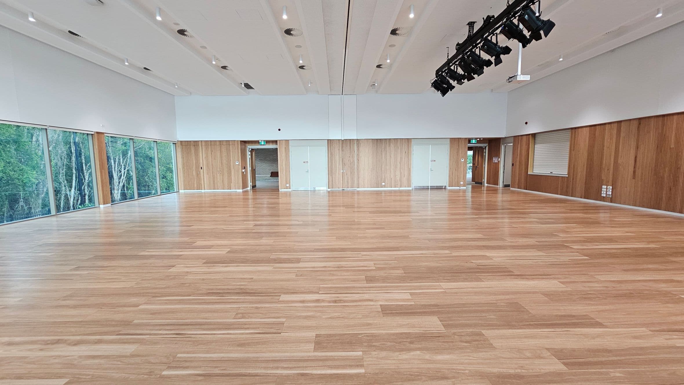 A spacious room with a light wooden floor and a high ceiling. On the left, large windows offer a view of green trees outside. The back wall features light wood panelling with several doors and an exit sign. On the right, a service window is set into the wall. Overhead, a row of black stage lights is suspended from the ceiling. The room is well-lit with recessed lighting.