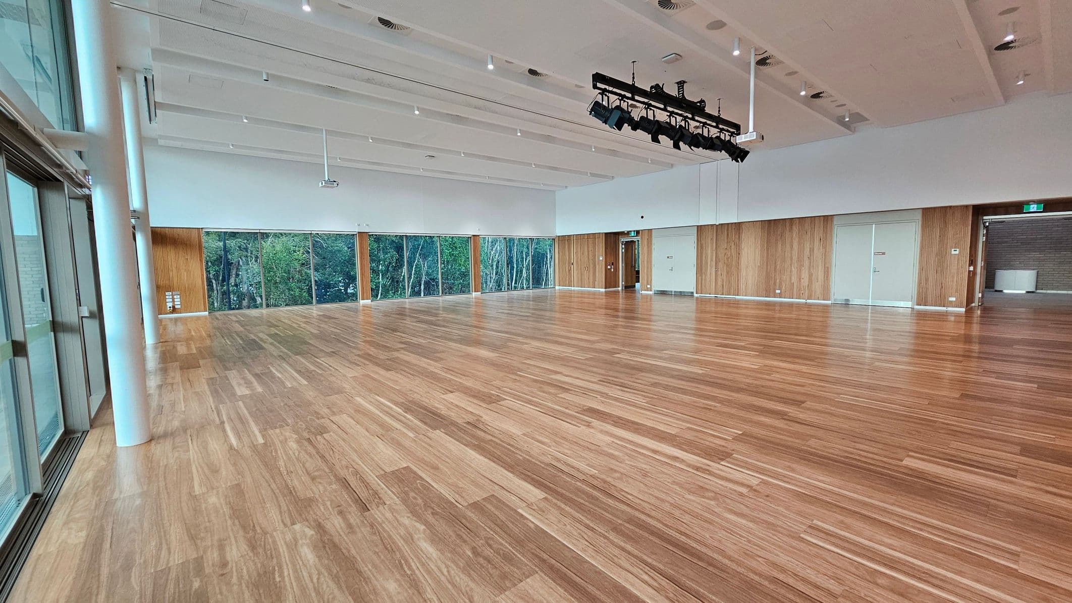 A spacious room with a polished wooden floor and a high white ceiling featuring recessed lighting and a mounted lighting rig. The left side has large glass windows with views of trees outside. The back wall is lined with wooden panels and has several doors, including a glass door and an emergency exit. The right side has a brick wall section with a counter. The room is well-lit and open, suitable for events or gatherings.