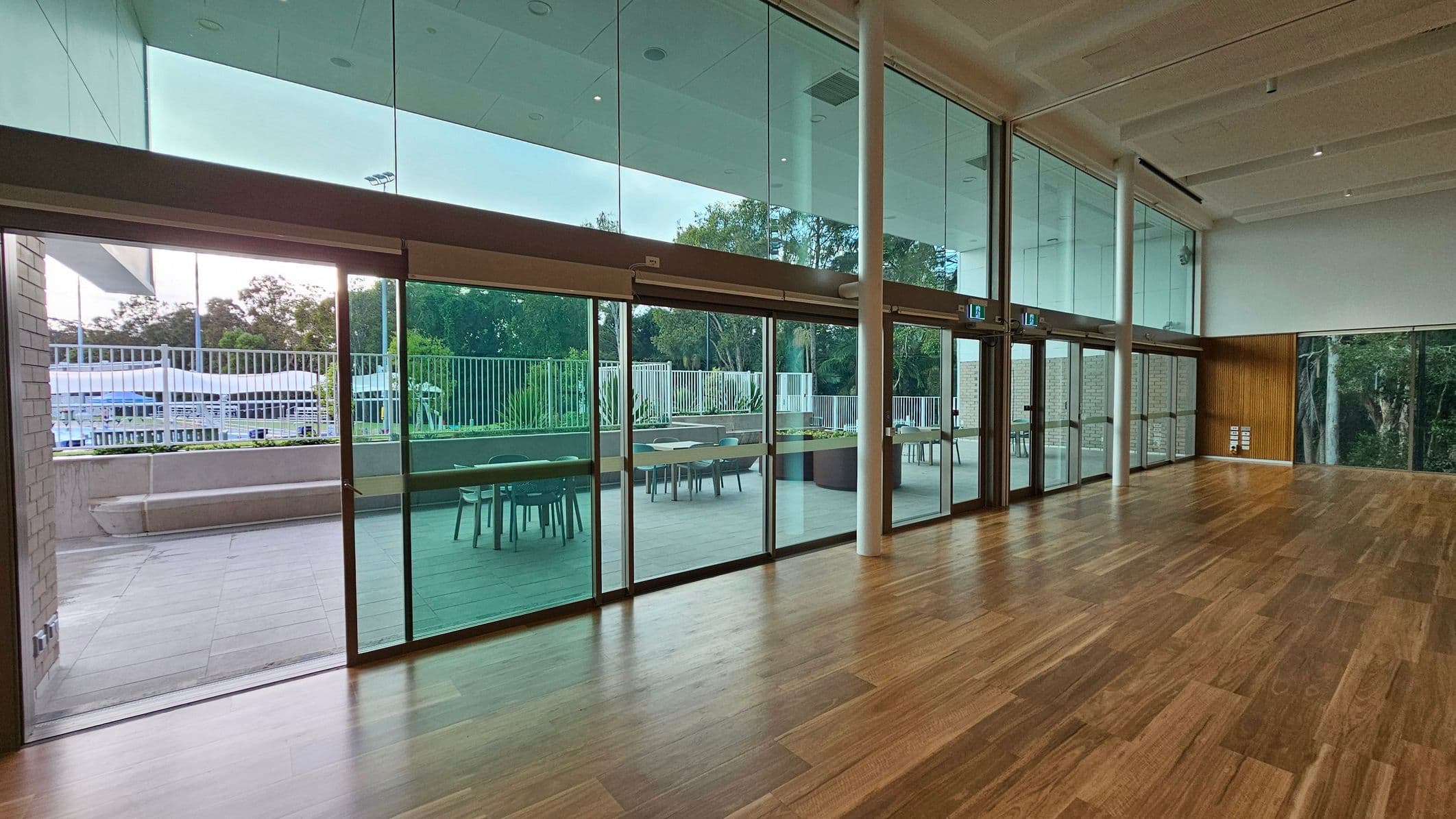A spacious indoor area with a wooden floor and a high ceiling featuring recessed lighting. The right side has large glass doors leading to an outdoor patio with tables and chairs. The patio is bordered by a white fence, and beyond it, there are trees and greenery. The left side shows more of the outdoor area, including a concrete bench. The ceiling inside is white with visible beams.
