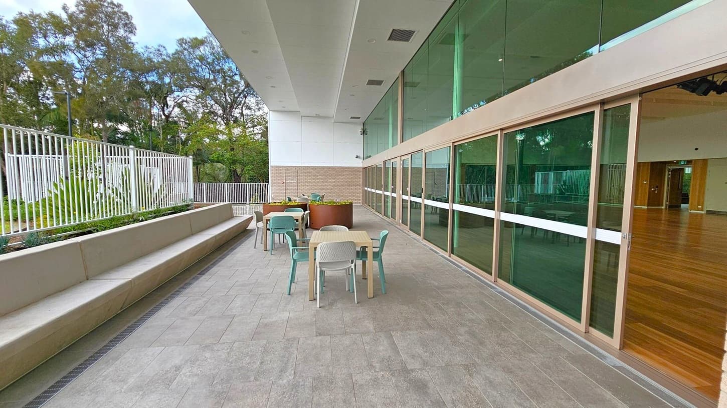 A spacious outdoor patio area with grey tiled flooring. On the left, there's a long bench with a white metal fence and greenery behind it. In the centre, several tables with chairs are arranged, and a large planter is visible. On the right, a building with large glass windows and wooden frames opens into an interior space with wooden flooring. The ceiling is white with recessed lighting. Trees are visible in the background.