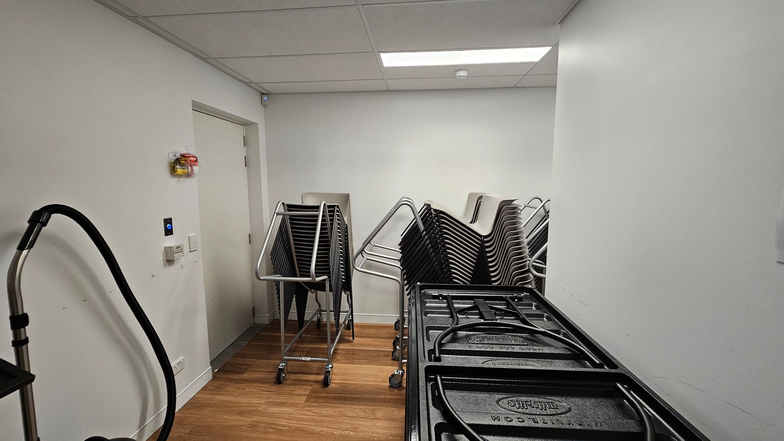 A storage room with a wooden floor and a white ceiling with fluorescent lighting. On the left, a vacuum cleaner with a black hose is visible. In the centre, there are stacks of black chairs on metal trolleys. On the right, there are large black plastic containers with handles. A closed door is on the left wall, with a small control panel and a fire alarm above it. The walls are plain white.