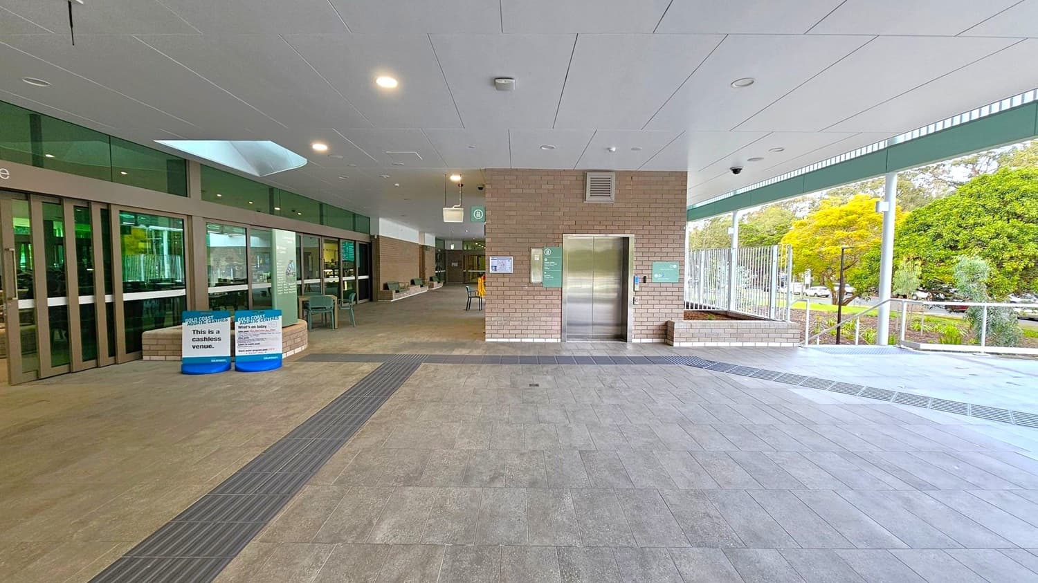 A covered outdoor area with a tiled floor and a white panelled ceiling. On the left, there are large glass doors with signs indicating a cashless venue. In the centre, a brick wall features a lift with signage and a control panel. To the right, a glass barrier overlooks a landscaped area with trees and a pathway. Several chairs and tables are scattered around, and there are overhead lights and a security camera.