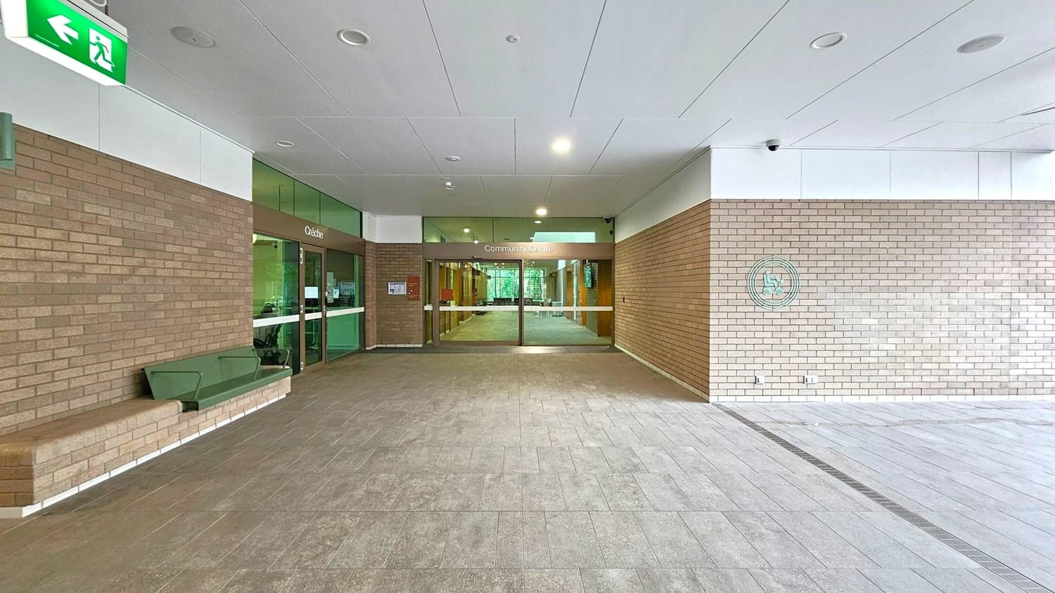 A spacious entrance area with a tiled floor and a ceiling with recessed lights. On the left, there's a brown brick wall with a green bench and a green exit sign above. In the centre, glass doors lead to a "Community Centre" with a crèche to the left. The right side features a light brown brick wall with a circular emblem. The area is well-lit and open, with a modern design.