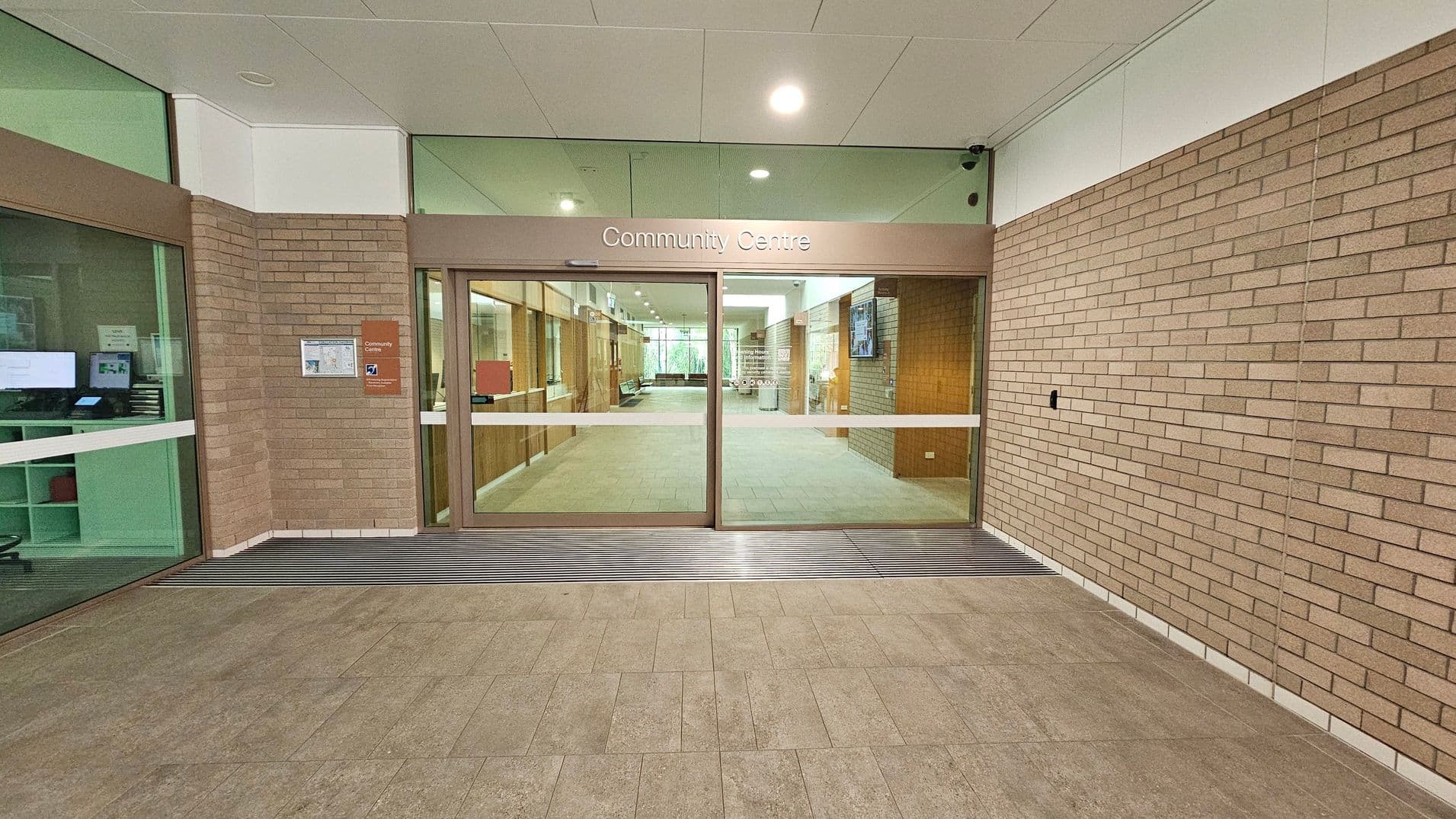 Entrance to a community centre with glass sliding doors in the centre, framed by brown brick walls. Above the doors, a sign reads "Community Centre". To the left, a window reveals a room with computer screens and shelves. The floor is tiled in a light brown pattern, and the ceiling is white with recessed lighting. Inside, a hallway extends with more brick walls and a glimpse of seating and notice boards.