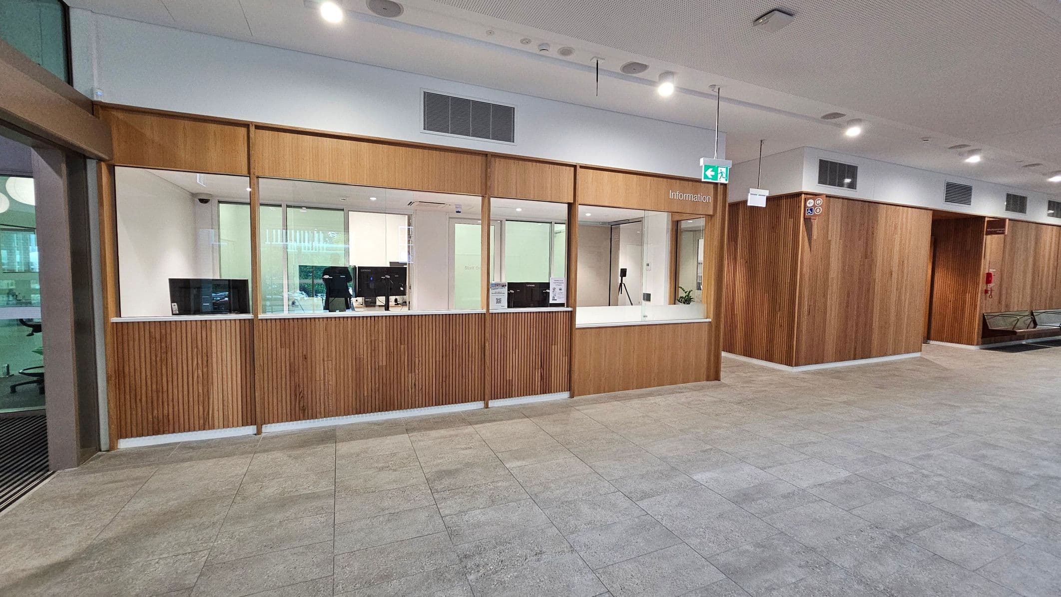 A modern interior with a wooden-panelled information desk in the centre, featuring multiple computer monitors. The desk is enclosed with glass partitions. To the right, a wooden wall with signage and a small seating area with black chairs. The floor is tiled in grey, and the ceiling is white with recessed lighting and ventilation panels. An exit sign hangs above the desk area.