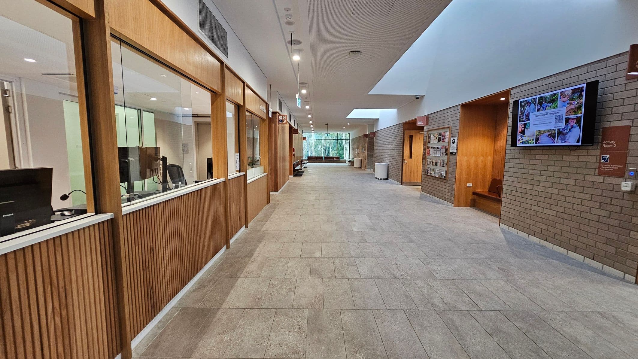 A corridor with a tiled floor and a ceiling with recessed lighting. On the left, there are wooden-panelled windows with office equipment visible inside. On the right, a brick wall features a digital screen displaying images and text, along with a sign for "Activity Room 2". Further down, there are brochures and a wooden bench. At the end of the corridor, large windows provide a view of trees outside.