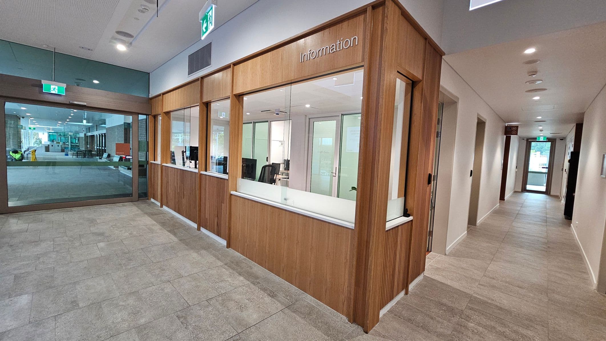 A modern interior with a tiled floor and a ceiling with recessed lighting. On the left, there's a large glass entrance with an exit sign above. In the centre, a wooden information desk with glass windows and a sign reading "Information" is visible. Behind the desk, there are office chairs and computers. On the right, a corridor with several doors leads to another glass exit door, also with an exit sign above.