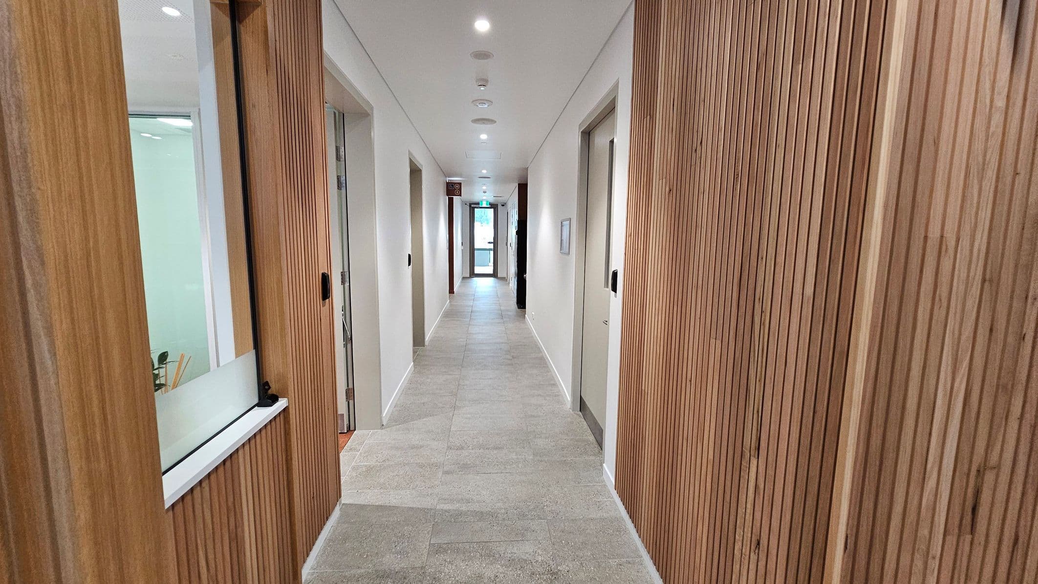 A corridor with light grey tiled flooring and white walls. The left and right sides feature wooden panelling with vertical grooves. Several doors are visible on both sides, with glass panels in some. Ceiling lights are evenly spaced along the corridor. At the end, a glass door leads outside, allowing natural light to enter.