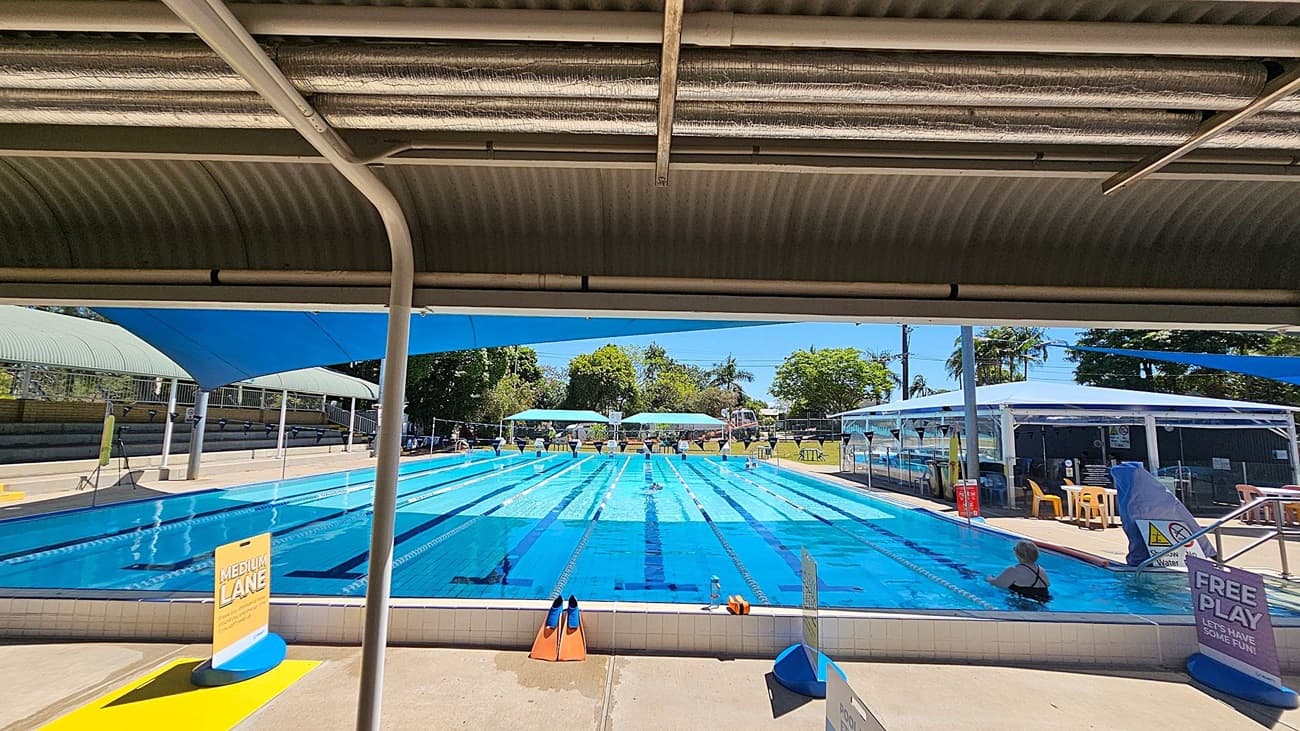 Outdoor swimming pool with multiple lanes, covered by blue shade sails. On the left, there is a concrete walkway and a tall light pole. In the middle, the pool has clear blue water with lane markers and signs indicating different lane speeds. On the right, a covered walkway with metal poles runs parallel to the pool. In the background, there are trees, a grassy area with tables and chairs, and a building with a corrugated roof.