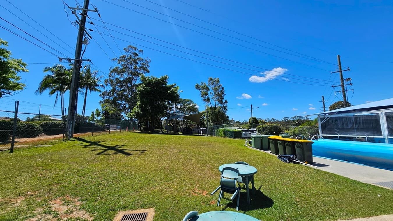 A grassy area with a few scattered patches of dirt. On the left, there is a chain-link fence with trees and power lines running above it. In the middle, there are several tall trees providing shade. On the right, there are multiple rubbish bins lined up next to a structure with a transparent cover. In the foreground, there are a few green plastic tables and chairs.