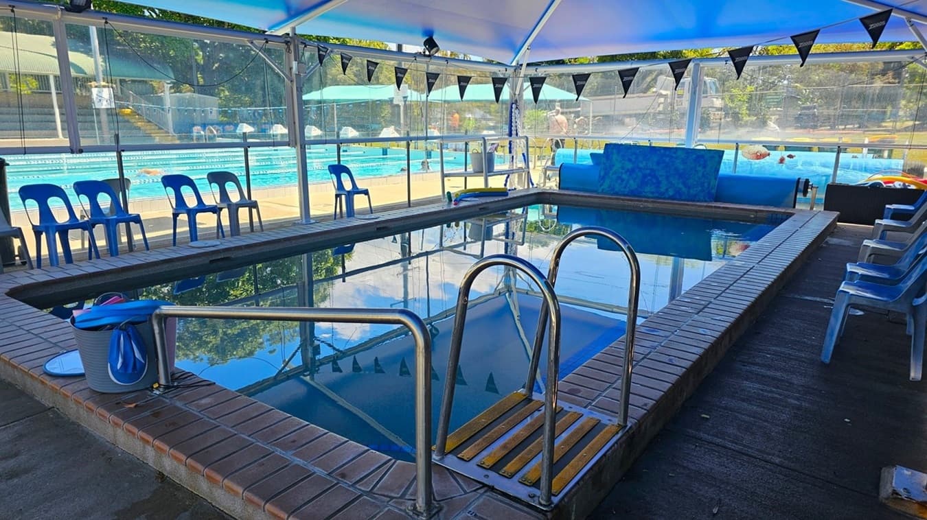 Indoor swimming pool with a tiled edge and clear water. On the left, there are blue plastic chairs lined up along the poolside. On the right, more blue plastic chairs are arranged similarly. In the foreground, a bucket filled with blue swimming equipment is placed near the pool's edge. The background shows a larger outdoor pool area with people swimming and a grassy area beyond. The ceiling is covered with a transparent material, allowing natural light to illuminate the space.