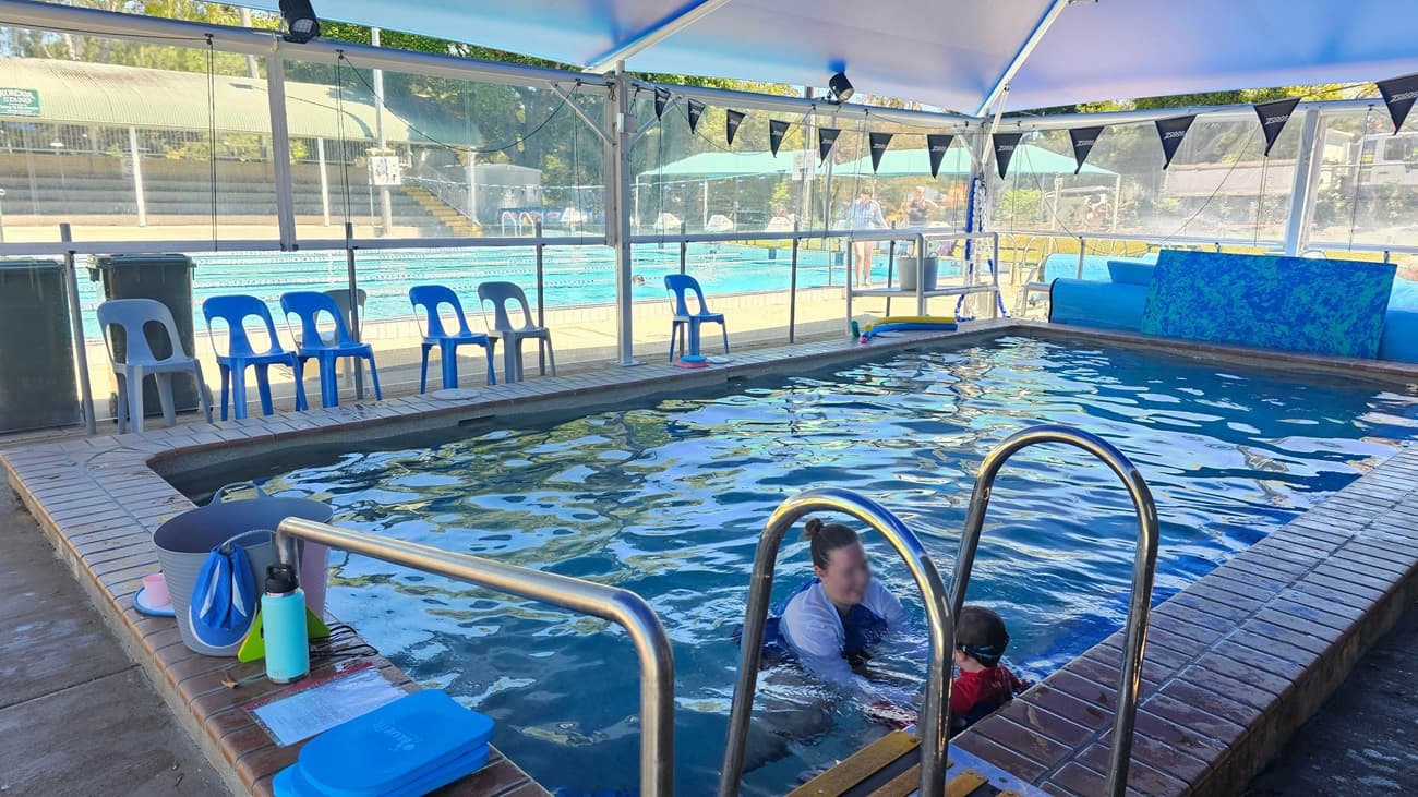 A small, rectangular indoor pool with a metal ladder on the right side. The pool is surrounded by a brick edge. Blue plastic chairs are lined up along the left side of the pool. In the background, there is a larger outdoor pool with lane dividers and more blue chairs. A transparent barrier separates the indoor and outdoor areas. The ceiling is covered with a blue canopy, and the floor is concrete. A bucket with swimming gear is placed near the pool's edge.