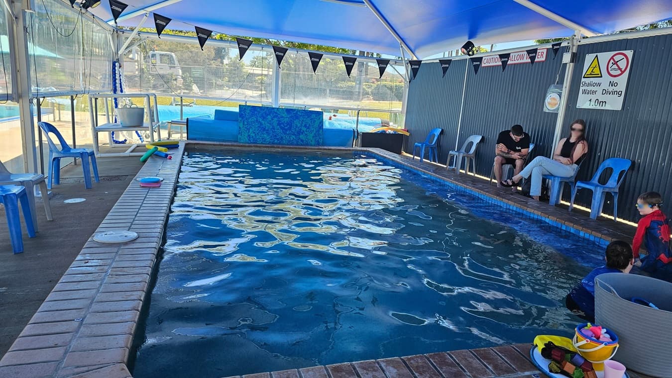 A small, rectangular indoor pool with a metal ladder on the left side. The pool is surrounded by a brick edge. Blue plastic chairs are lined up along the left side of the pool. In the background, there is a larger outdoor pool with lane dividers and more blue chairs. A transparent barrier separates the indoor and outdoor areas. The ceiling is covered with a blue canopy, and the floor is concrete. A bucket with swimming gear is placed near the pool's edge.